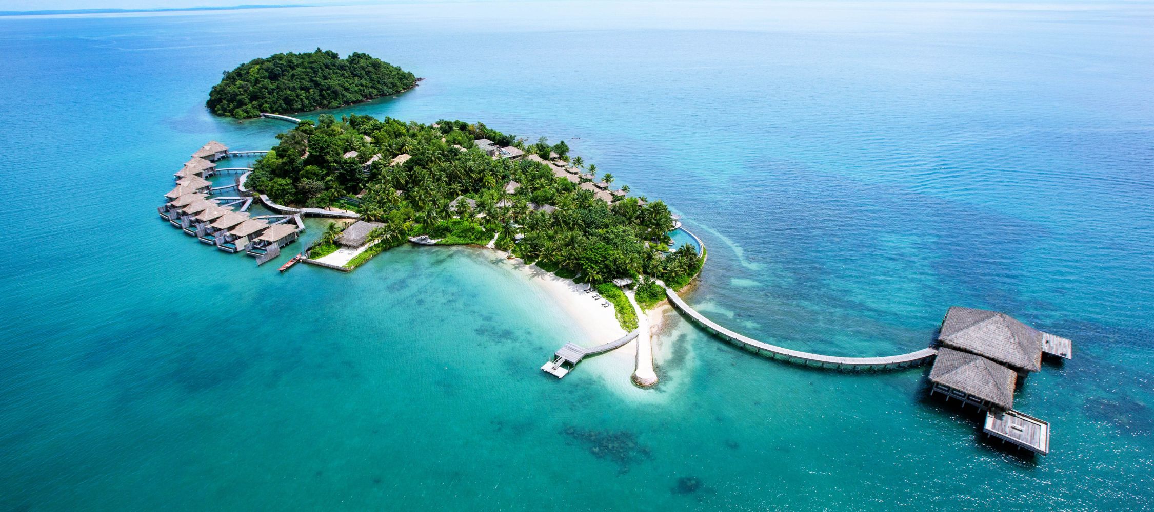 Aerial view of a tropical island resort featuring overwater bungalows, lush greenery, and clear blue water. The shoreline curves gently with sandy beaches, and a wooden walkway connects the main area to the bungalows.