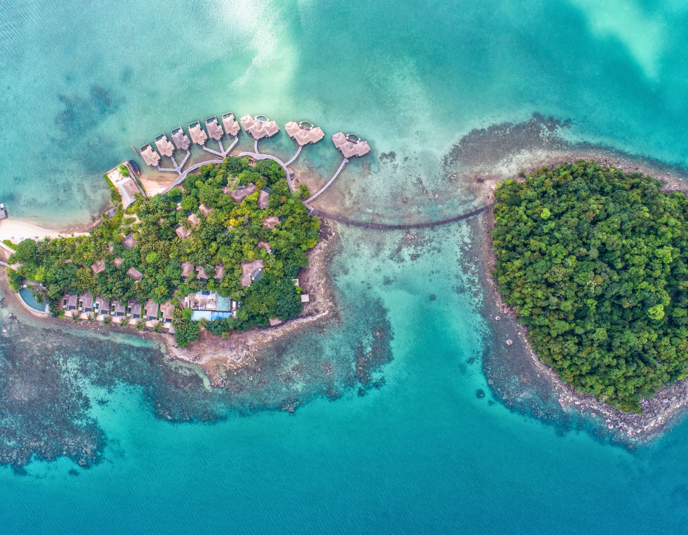 Aerial view of a tropical island resort with overwater bungalows connected by wooden walkways, surrounded by turquoise waters and lush greenery. A smaller, densely forested island is visible nearby.