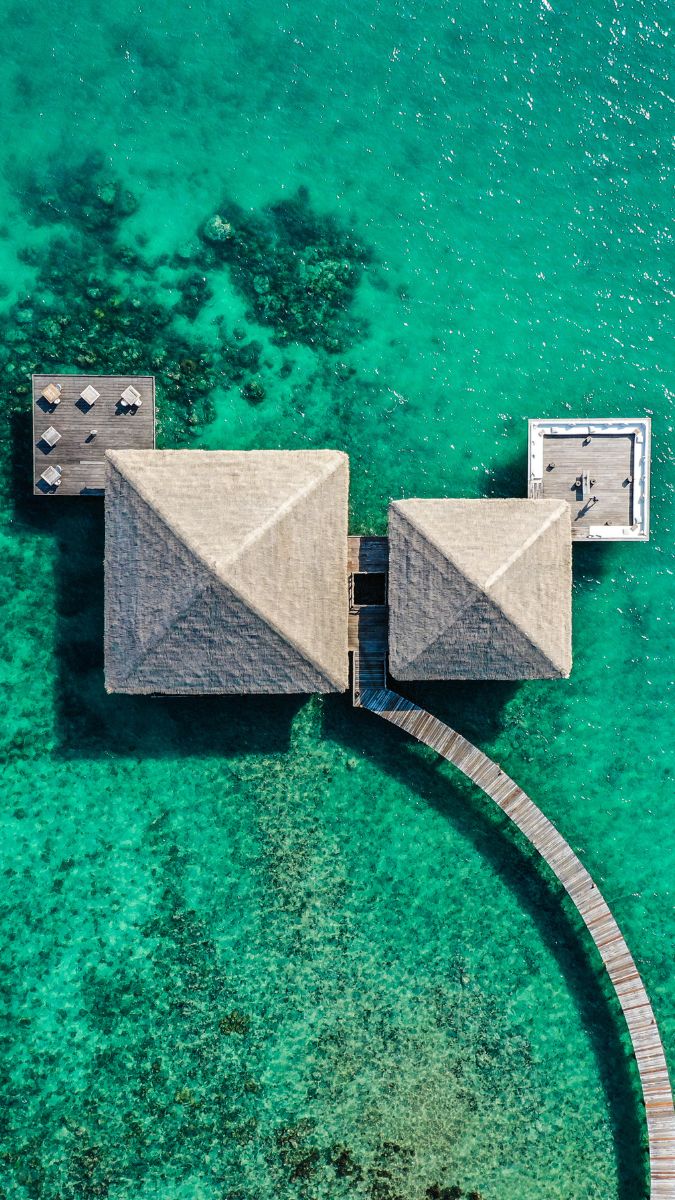 Overhead view of two thatched-roof huts connected by a wooden walkway, surrounded by crystal clear turquoise water. A deck area with lounge chairs is visible nearby.
