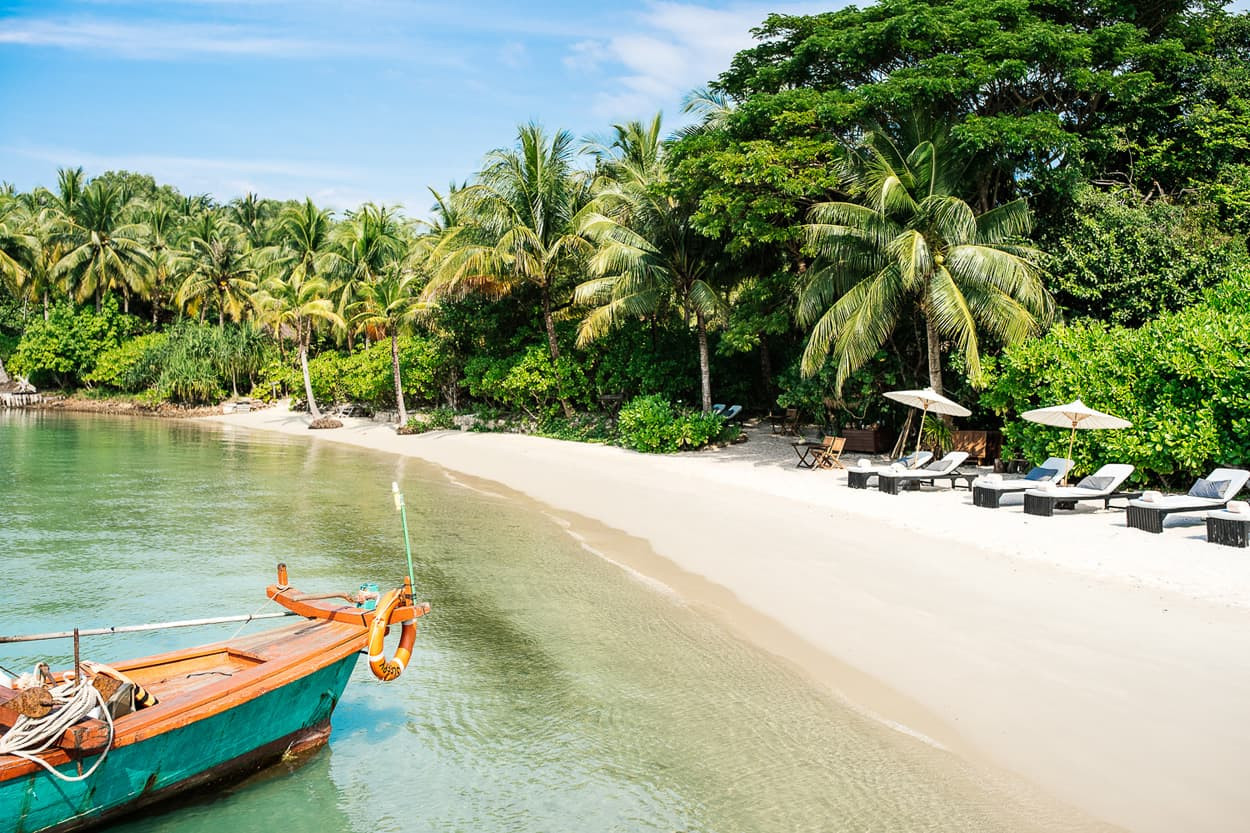 A tranquil beach scene with soft white sand and clear turquoise water. A colorful wooden boat is moored near the shore, surrounded by lush green palm trees. Sun loungers with umbrellas are set up along the beach under the shade of tropical foliage.