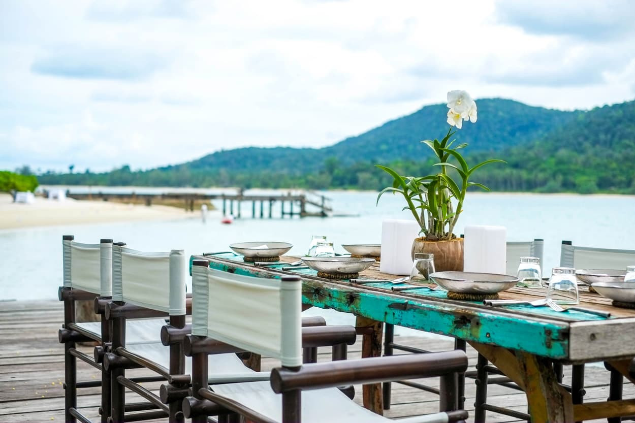A rustic dining table set for a meal, featuring white dishes and glassware, with a potted orchid centerpiece. In the background, a serene beach and calm water stretch out beneath a cloudy sky, framed by lush green hills.