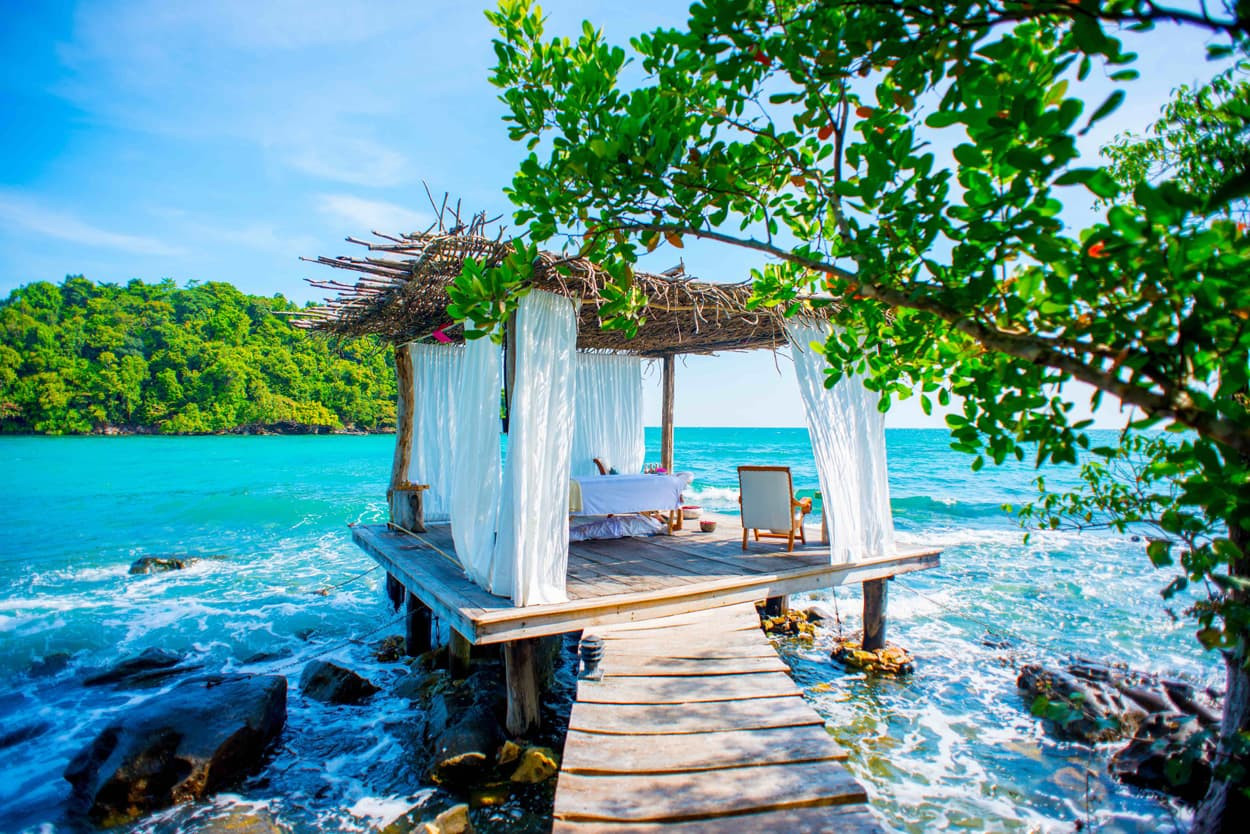 A wooden platform with a thatched roof and flowing white curtains extends over the turquoise ocean. Gentle waves lap against the structure, surrounded by lush greenery and a clear blue sky. A single chair and a massage table are set up on the platform, creating a serene escape.