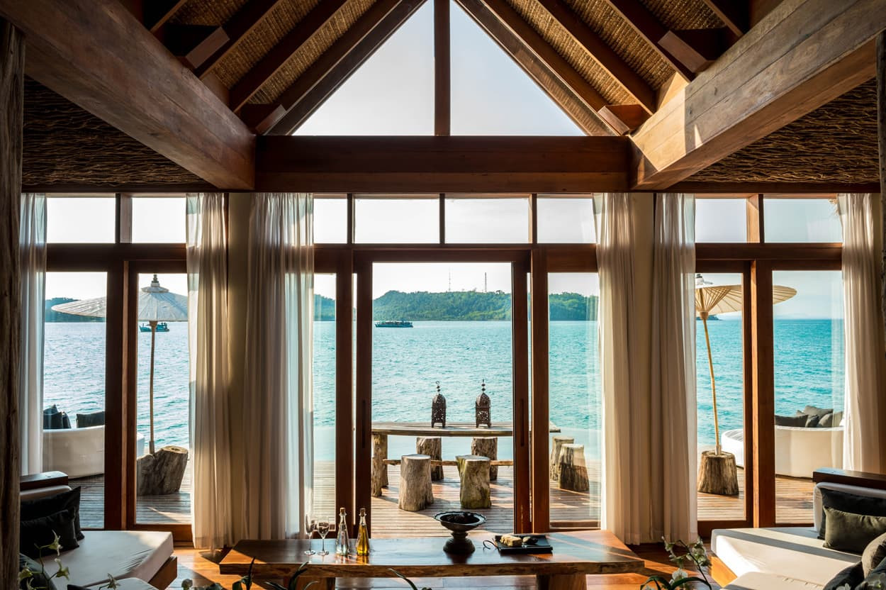 A luxurious beachfront villa with large windows overlooking the ocean. The interior features wooden beams and natural decor with a cozy seating area. Outside, a spacious deck has a dining table and parasols, framed by lush greenery and distant hills.