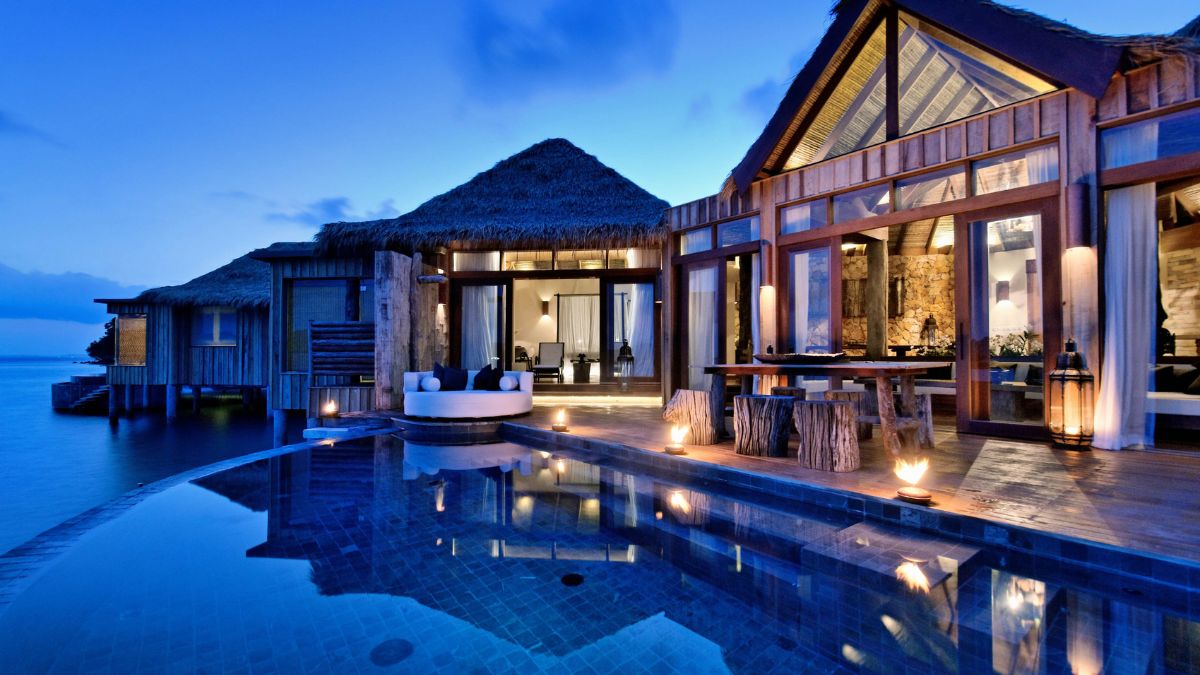 A luxurious overwater villa at dusk, featuring a thatched roof, large windows, and wooden decks. The villa is surrounded by calm water, with warm lights illuminating the pool area and pathways.