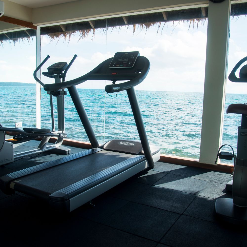 A treadmill in a gym with large glass windows overlooking the ocean. The calm water reflects the sky, creating a serene workout environment.