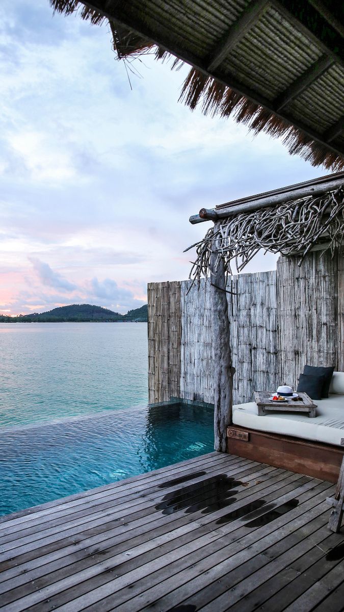 A tranquil view of a wooden deck leading to a serene infinity pool, overlooking calm waters. A thatched roof hut provides shade, and distant hills are visible under a pastel sky at sunset.