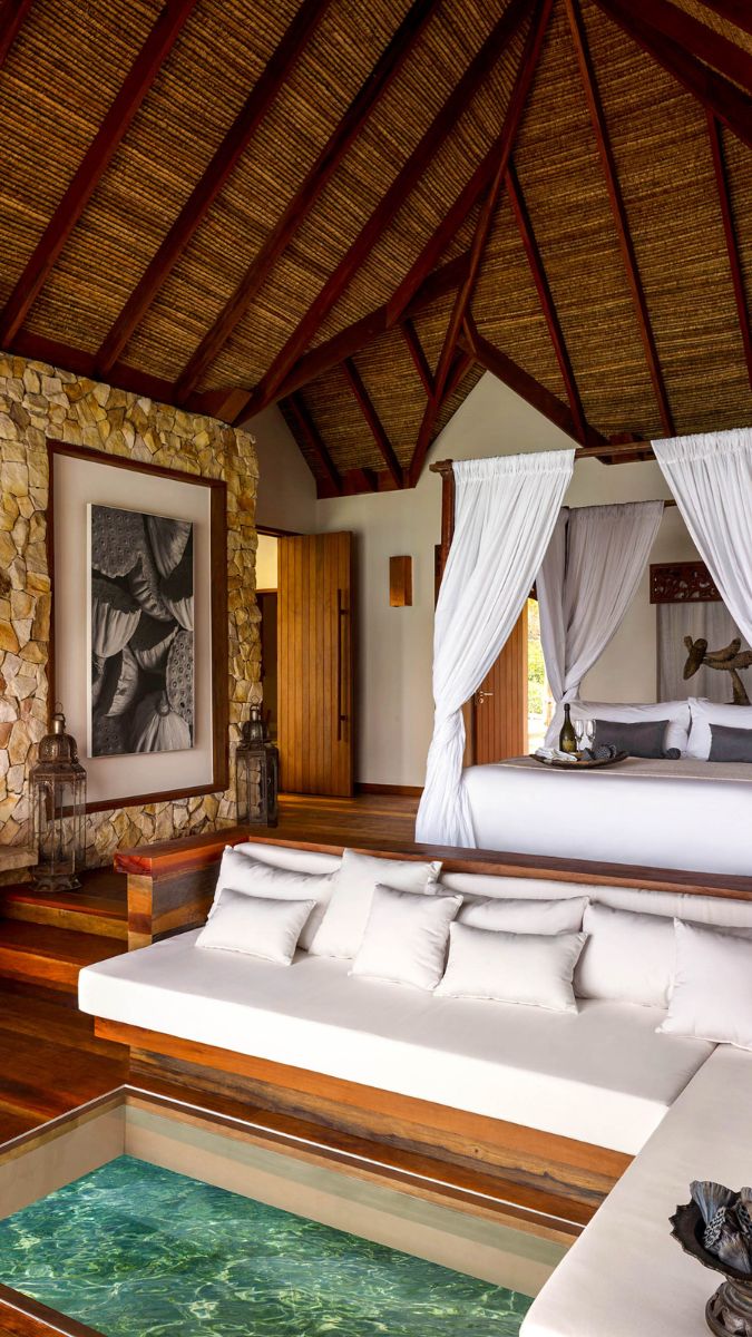 Luxurious hotel room featuring a thatched roof, wooden accents, and a serene seating area with white cushions. A glass floor panel reveals water beneath, and elegant curtains frame the bed area.