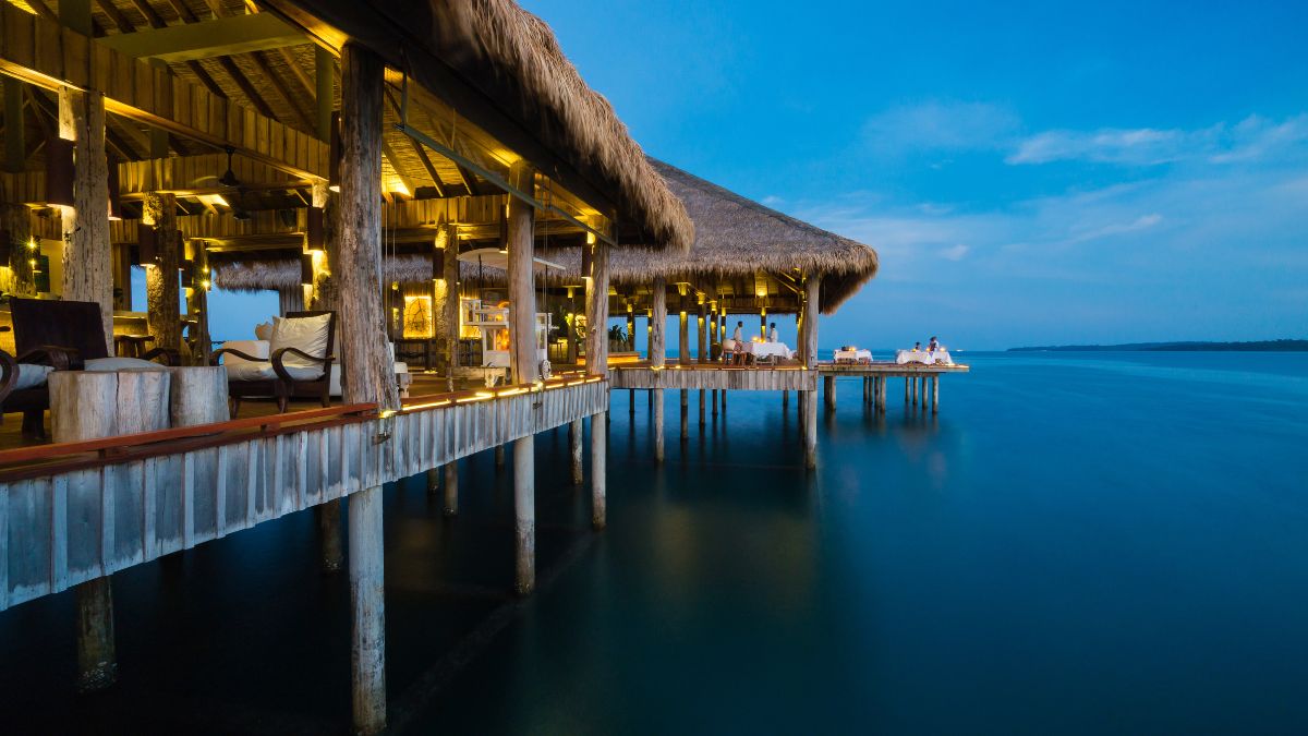 A luxurious wooden pavilion with a thatched roof, extending over calm waters at dusk. Soft lighting illuminates the seating area, and the serene blue sky reflects on the water's surface.