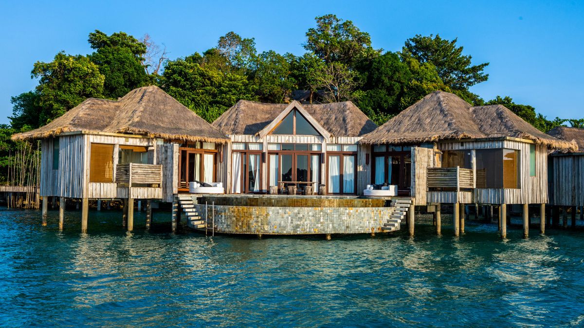 Luxury waterfront villas with thatched roofs are elevated on stilts over clear blue water, surrounded by lush greenery. A spacious deck extends from the center villa, featuring seating areas.