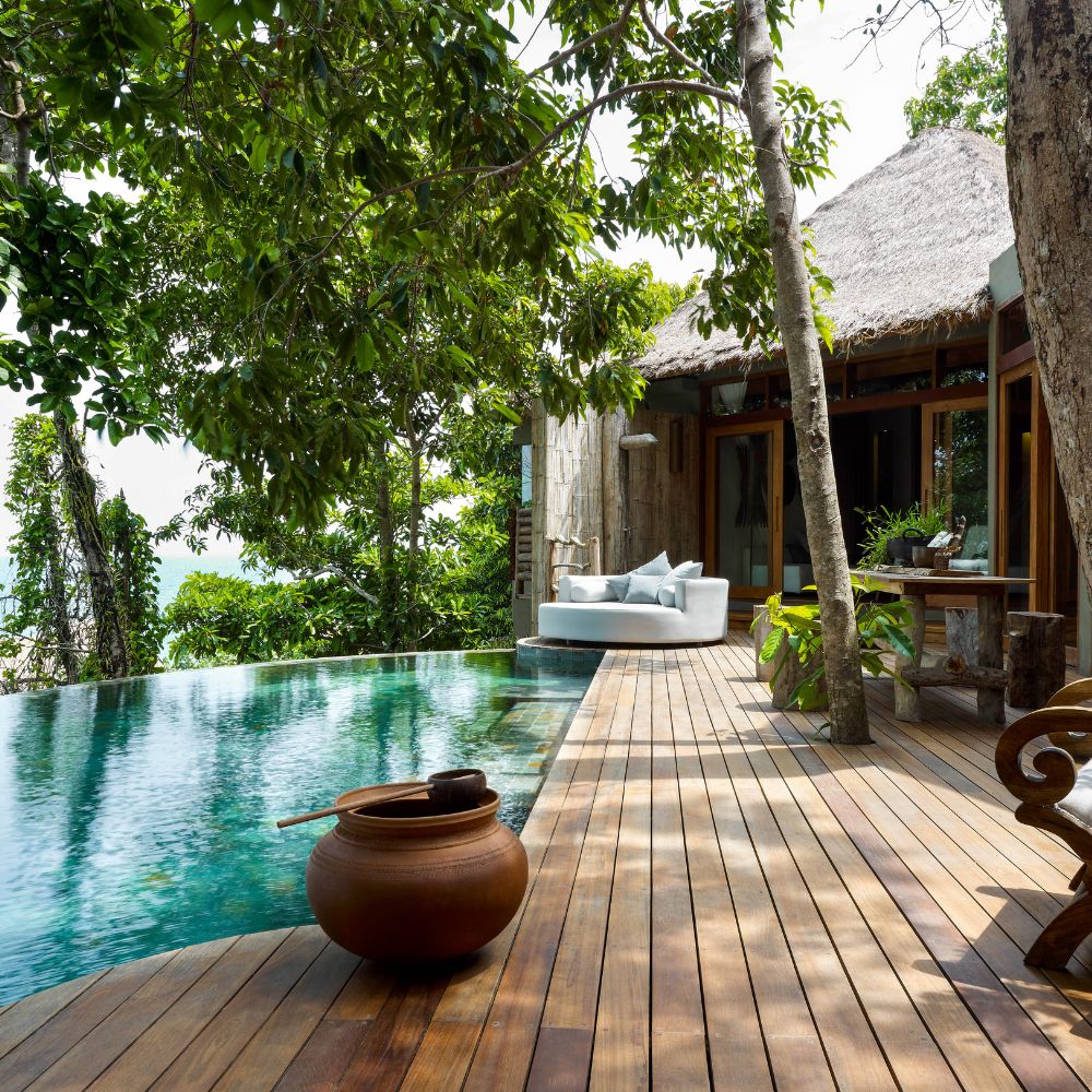 A serene outdoor space features a wooden deck leading to a tranquil infinity pool surrounded by lush greenery. A comfortable white sofa is positioned near the pool, alongside a decorative clay pot. The setting exudes a peaceful, tropical atmosphere.