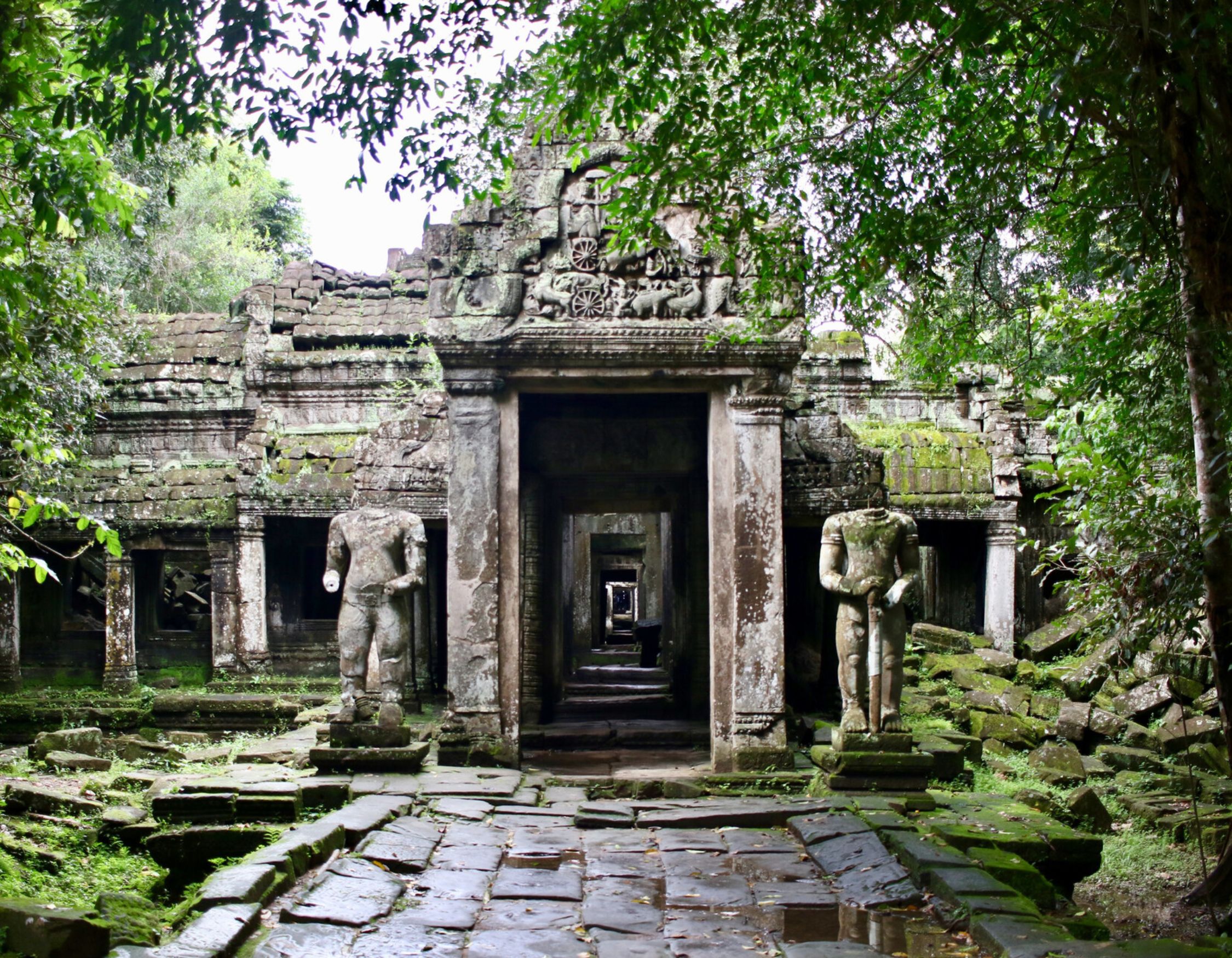Ancient stone temple ruins surrounded by lush greenery, featuring moss-covered walls and two weathered statues flanking the entrance. The pathway is lined with large stone slabs leading into the temple.