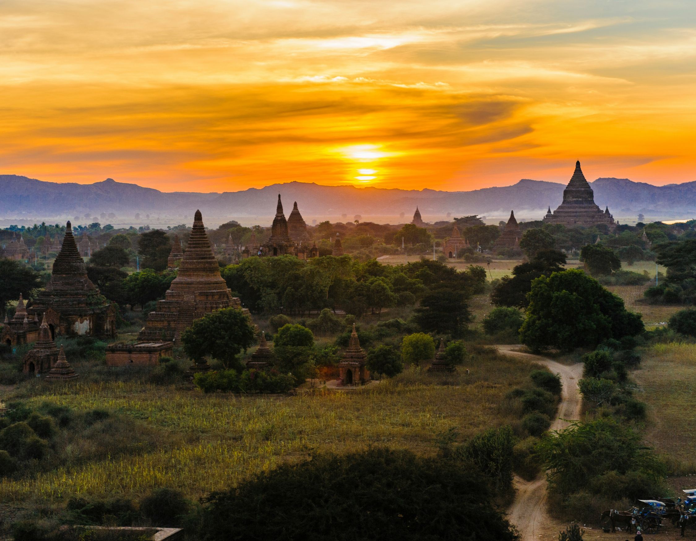A stunning sunset over ancient temples and pagodas in a lush landscape, with silhouettes of hills in the background. Vibrant orange and yellow hues fill the sky, creating a serene atmosphere.
