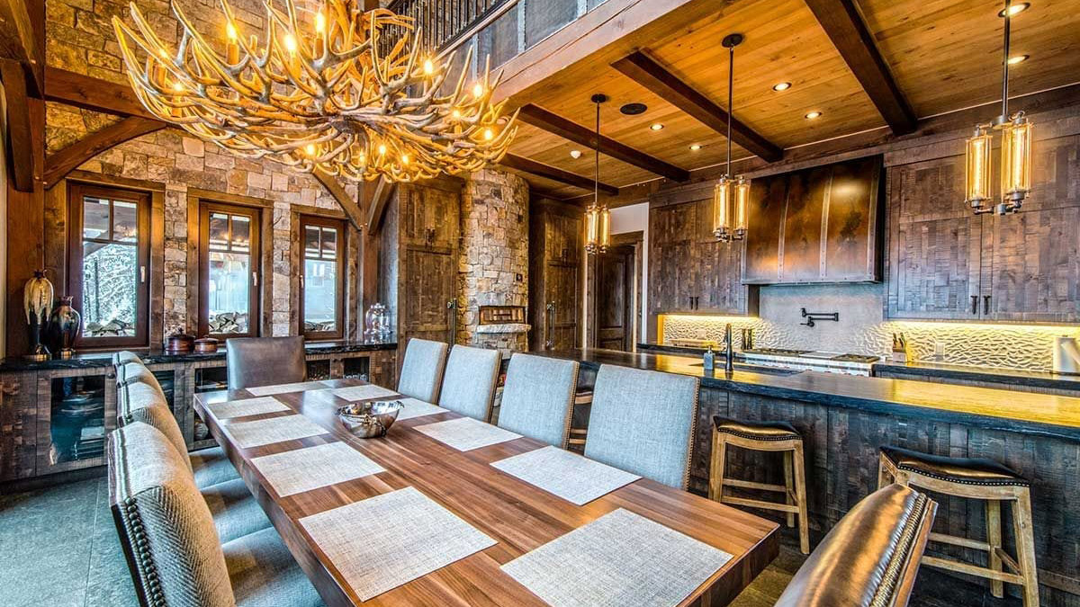 A rustic dining room with a large wooden table surrounded by comfortable gray chairs. An impressive antler chandelier hangs from the ceiling, illuminating the space. Stone and wood walls provide a warm, inviting atmosphere, complemented by modern light fixtures and a sleek kitchen area in the background.