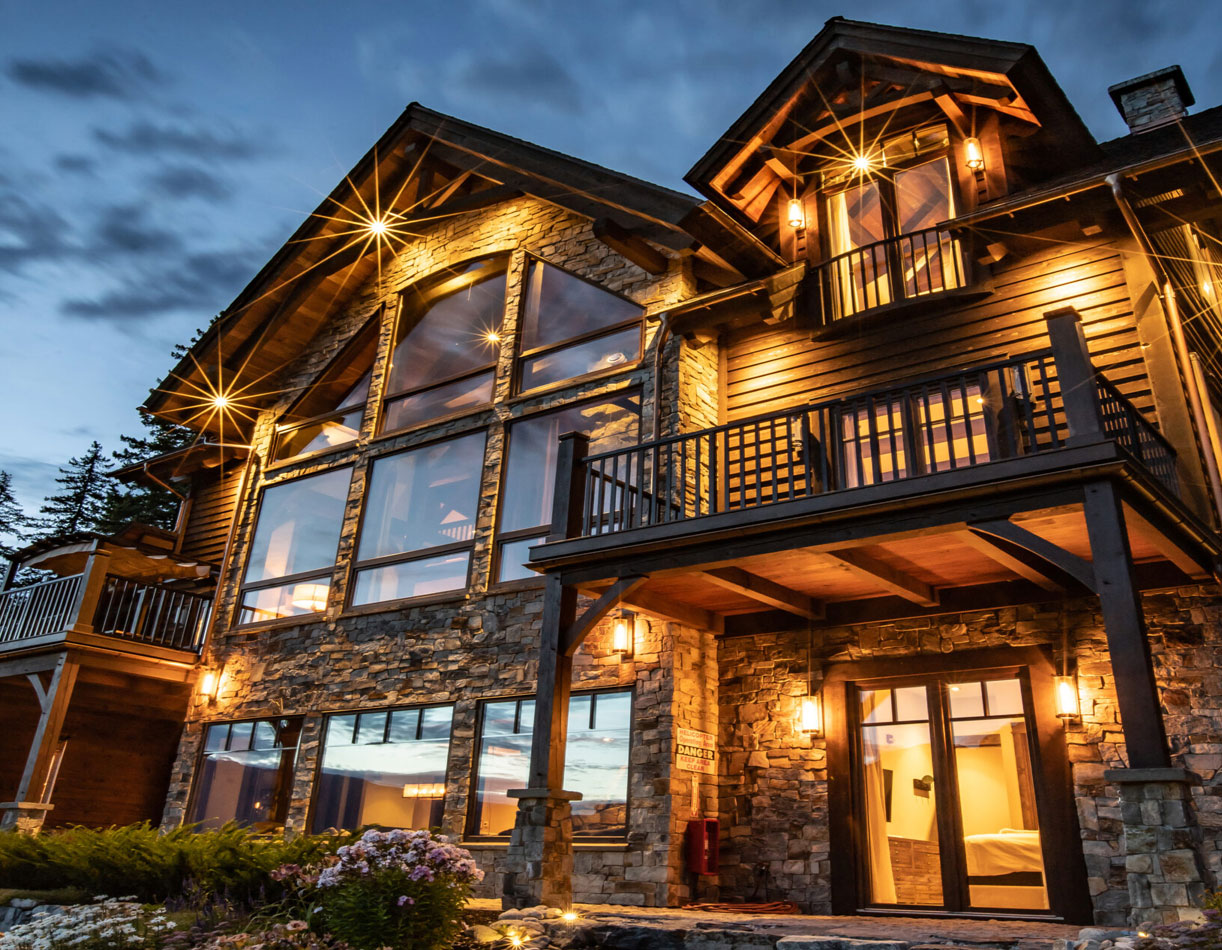 A modern chalet with large windows, stone exterior, and wooden balconies, illuminated by warm outdoor lights against a twilight sky.