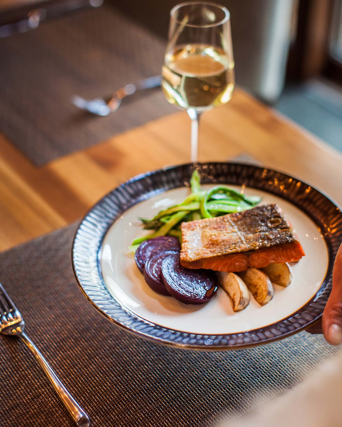 A beautifully plated dish featuring a piece of salmon alongside roasted potatoes, beetroot, and green beans. A glass of white wine is nearby on a wooden table.