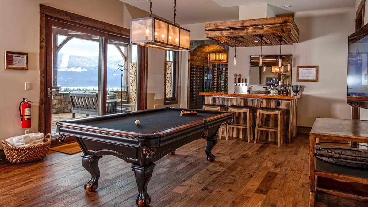 A beautifully designed room featuring a billiard table, wooden flooring, and a bar area with stools. Large windows offer a stunning view of mountains and nature.