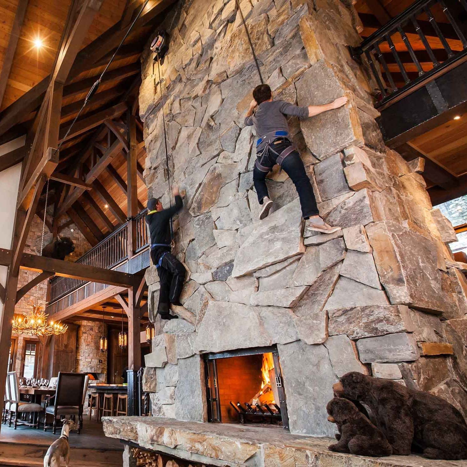 Two climbers ascend a large, rugged stone wall within a rustic, wooden-laden interior. A roaring fireplace sits at the base, and a small dog watches from nearby. Warm lighting adds to the cozy atmosphere.
