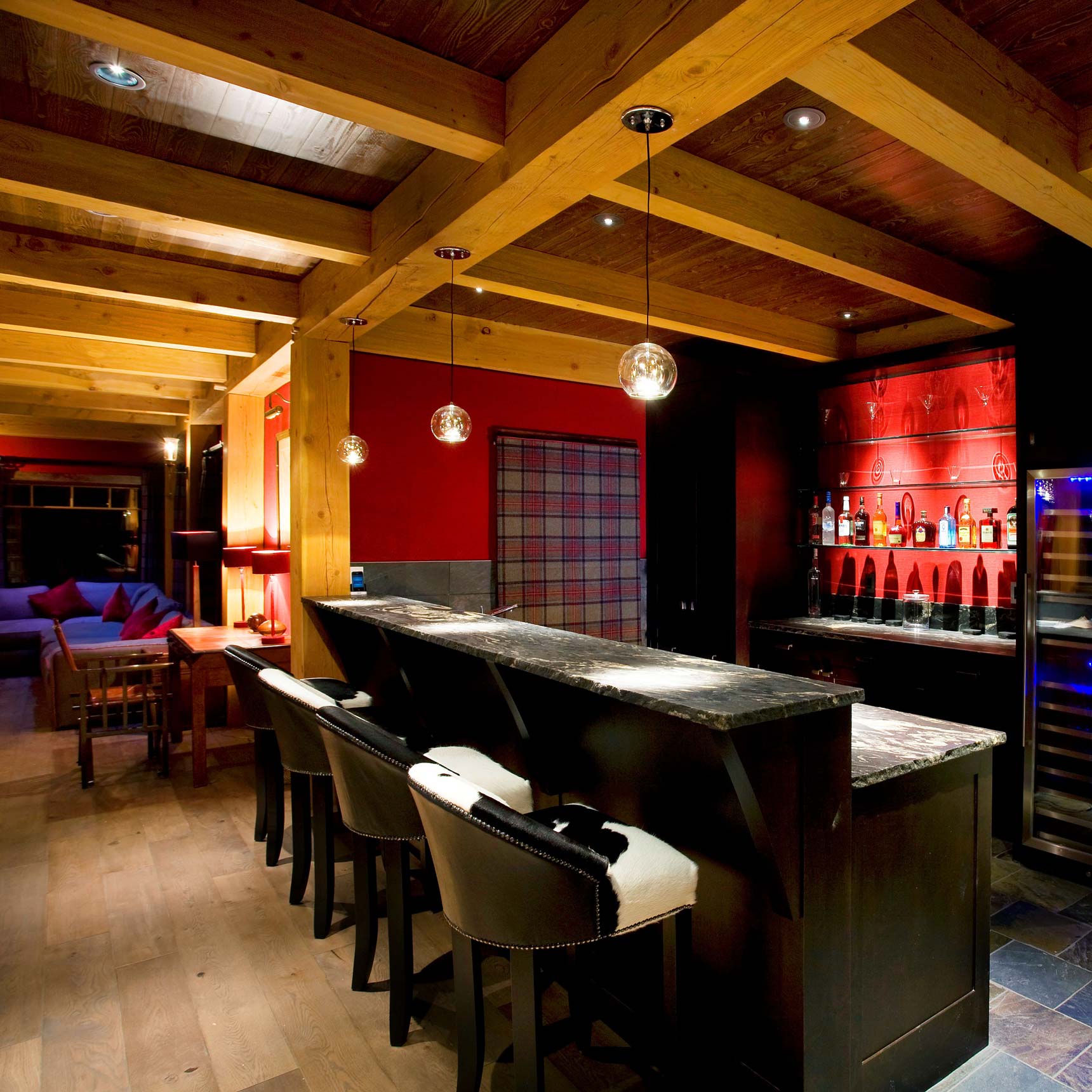 A modern home bar with a sleek black counter and high-backed chairs. Pendant lights hang above the bar, while a colorful wall with bottles of liquor is visible in the background. Exposed wooden beams and warm lighting create an inviting atmosphere.