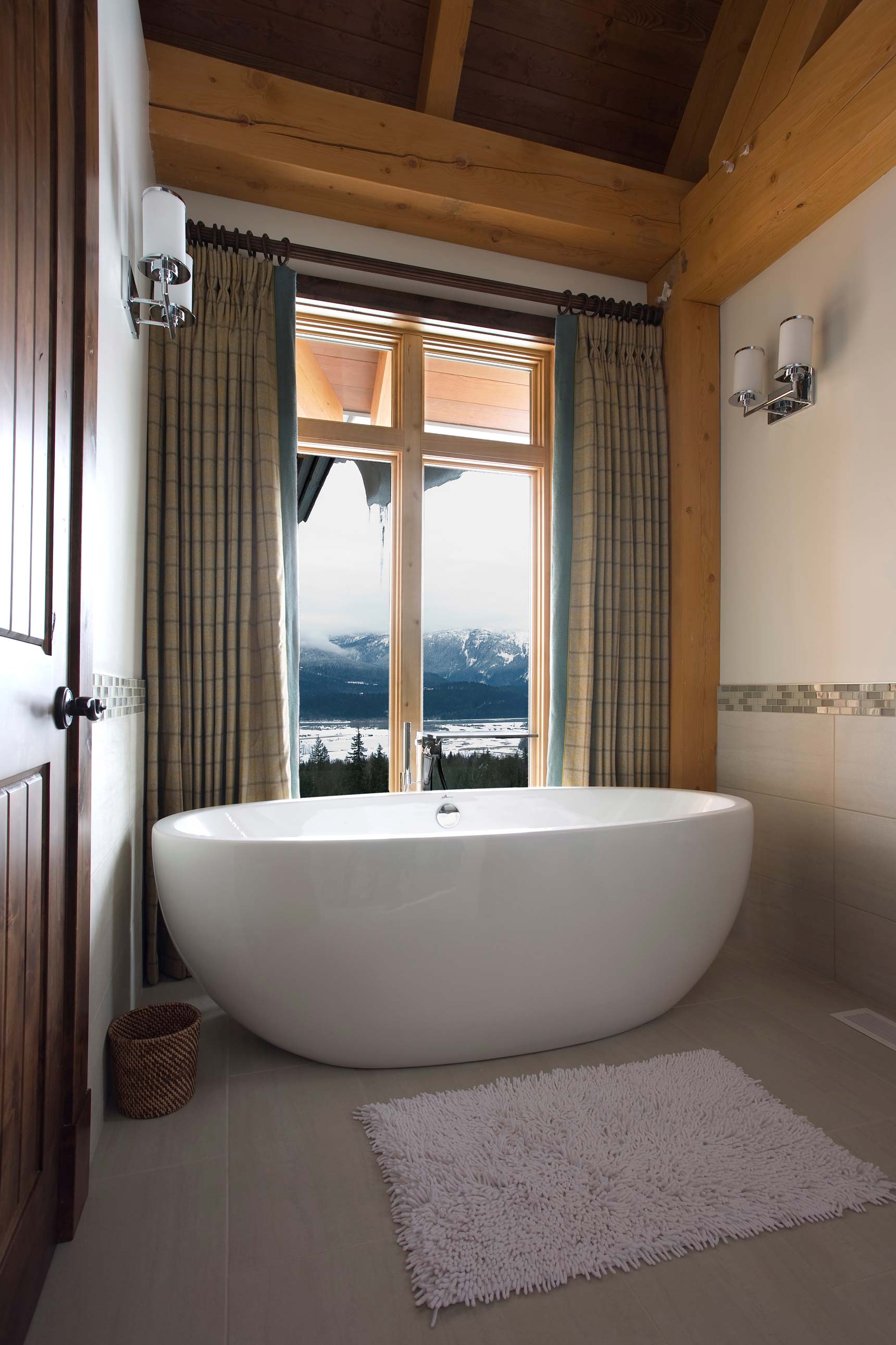 A modern bathroom featuring a freestanding white bathtub next to large windows with stunning mountain views. The space is adorned with wooden beams, neutral walls, and soft lighting, creating a serene atmosphere. A fluffy white rug sits on the floor beside the tub.