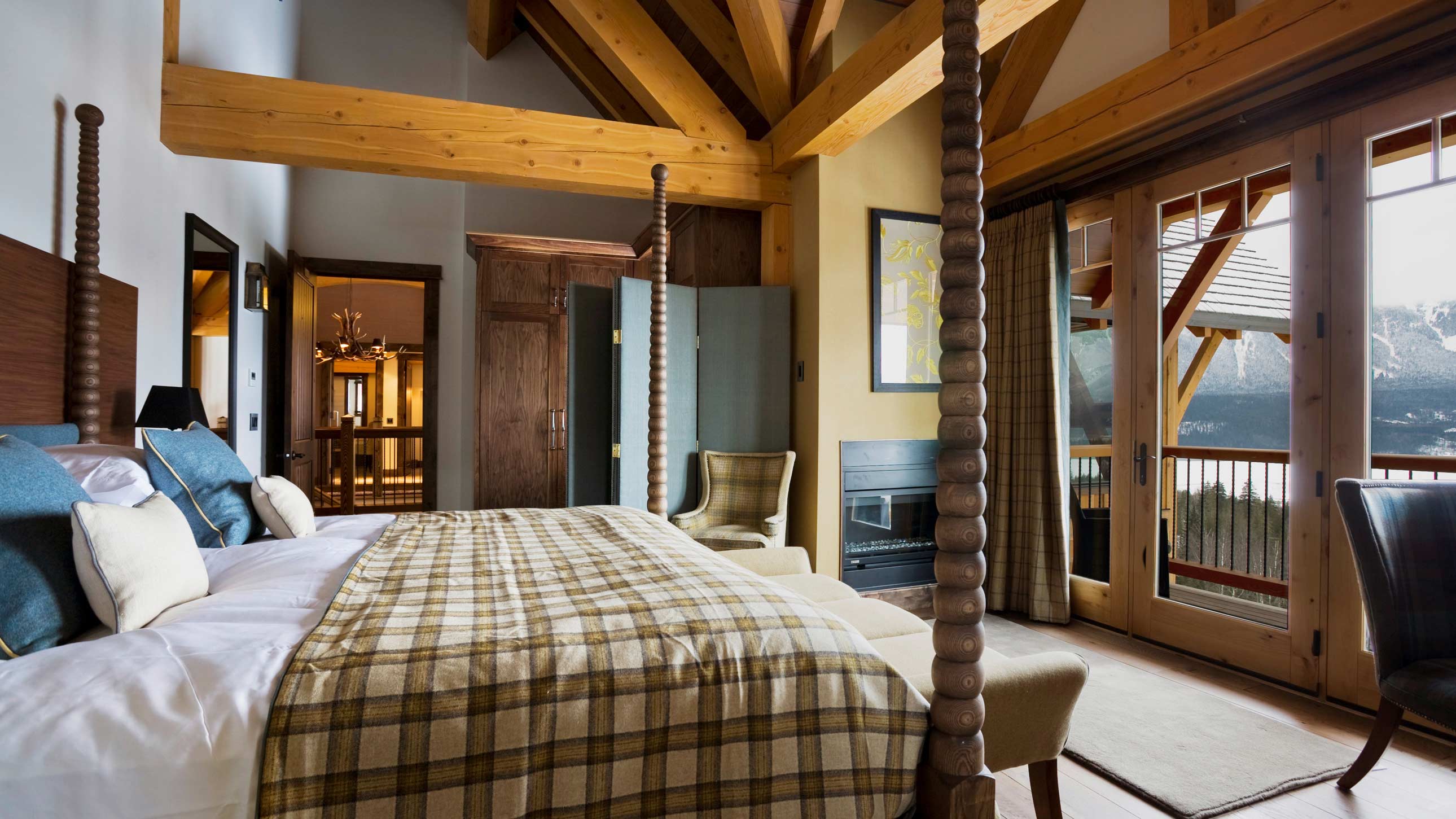A stylish, spacious bedroom featuring a wooden ceiling with exposed beams, a large bed adorned with a checkered throw, and elegant armchairs. Double doors open to a balcony with scenic views, while warm lighting creates a cozy atmosphere.