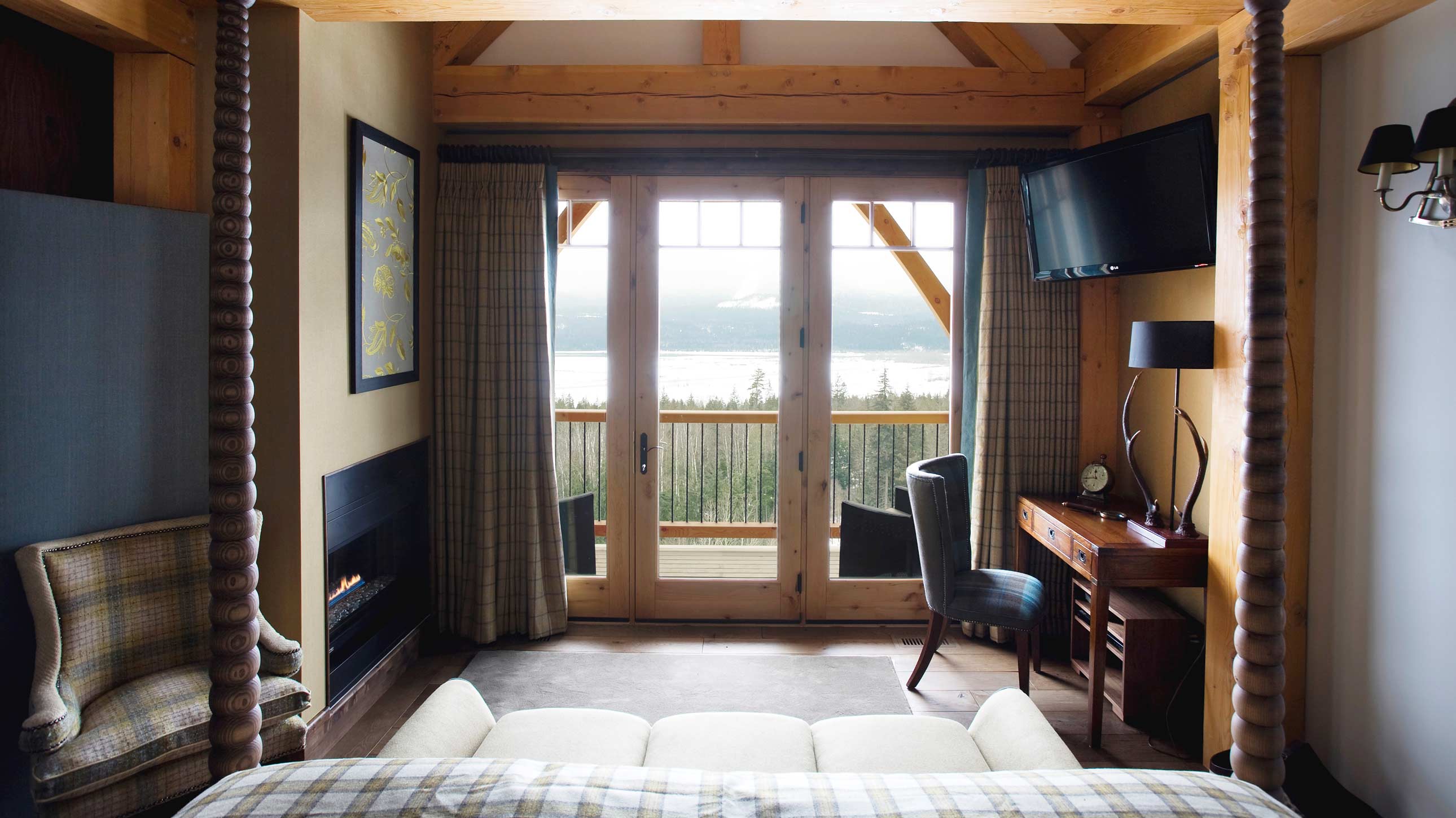 Cozy hotel room featuring a comfortable bed, a wooden balcony with a stunning view of a lake, and a desk with a chair. Large windows allow natural light, creating a warm atmosphere.