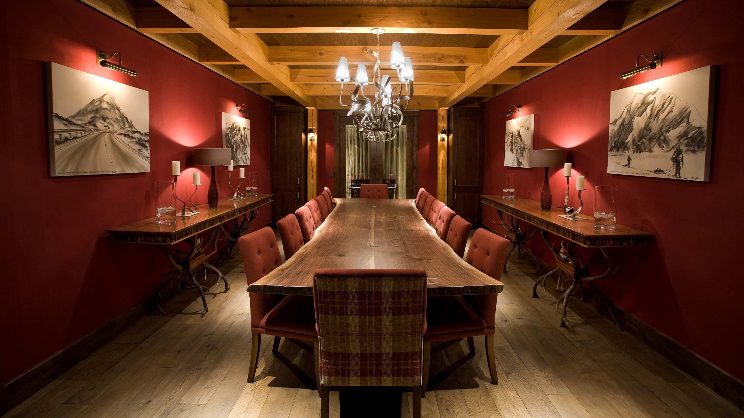 A rustic dining room featuring a long wooden table surrounded by red upholstered chairs. The walls are painted deep red, adorned with black and white mountain-themed artwork. A chandelier hangs from the wooden ceiling beams, and warm lighting from wall sconces adds an inviting ambiance.