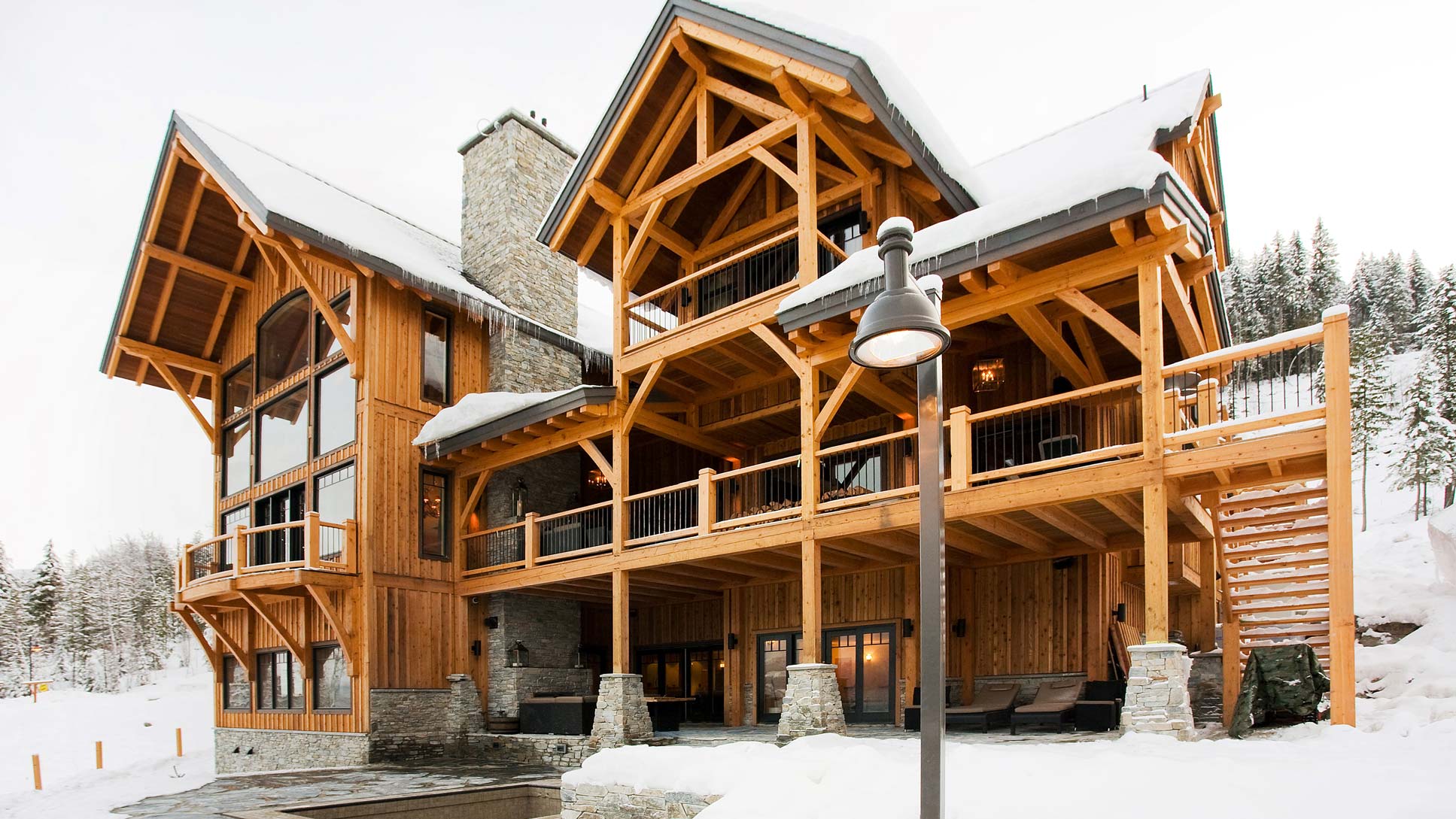 A large wooden lodge with a stone fireplace, featuring expansive windows and balconies. Surrounded by snow-covered landscapes and pine trees, it exudes a cozy, rustic charm.