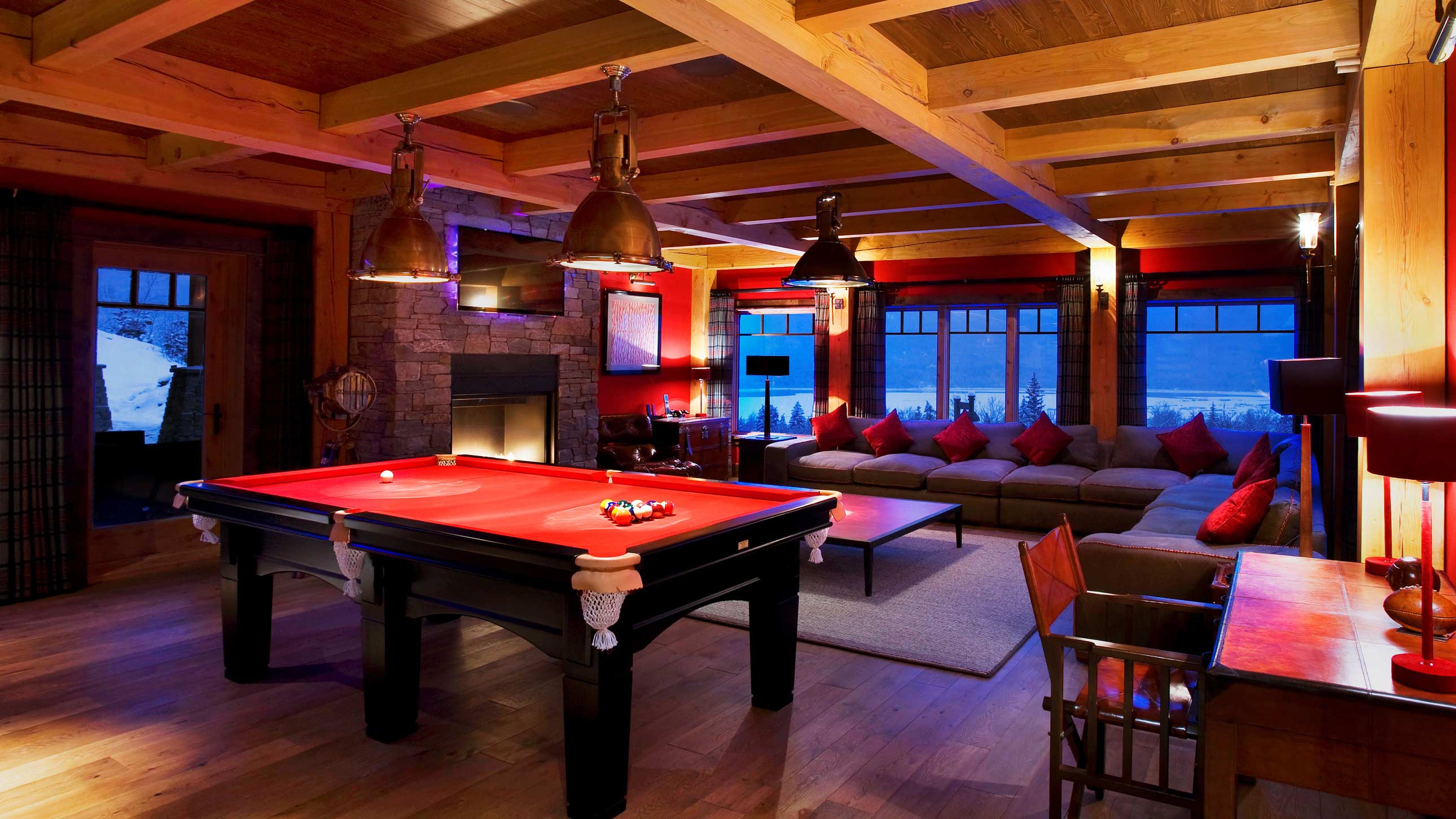 A cozy living room with a red pool table, wooden beams, and large windows showcasing a snowy landscape. The room features a comfortable sofa with cushions, warm lighting, and stone accents.