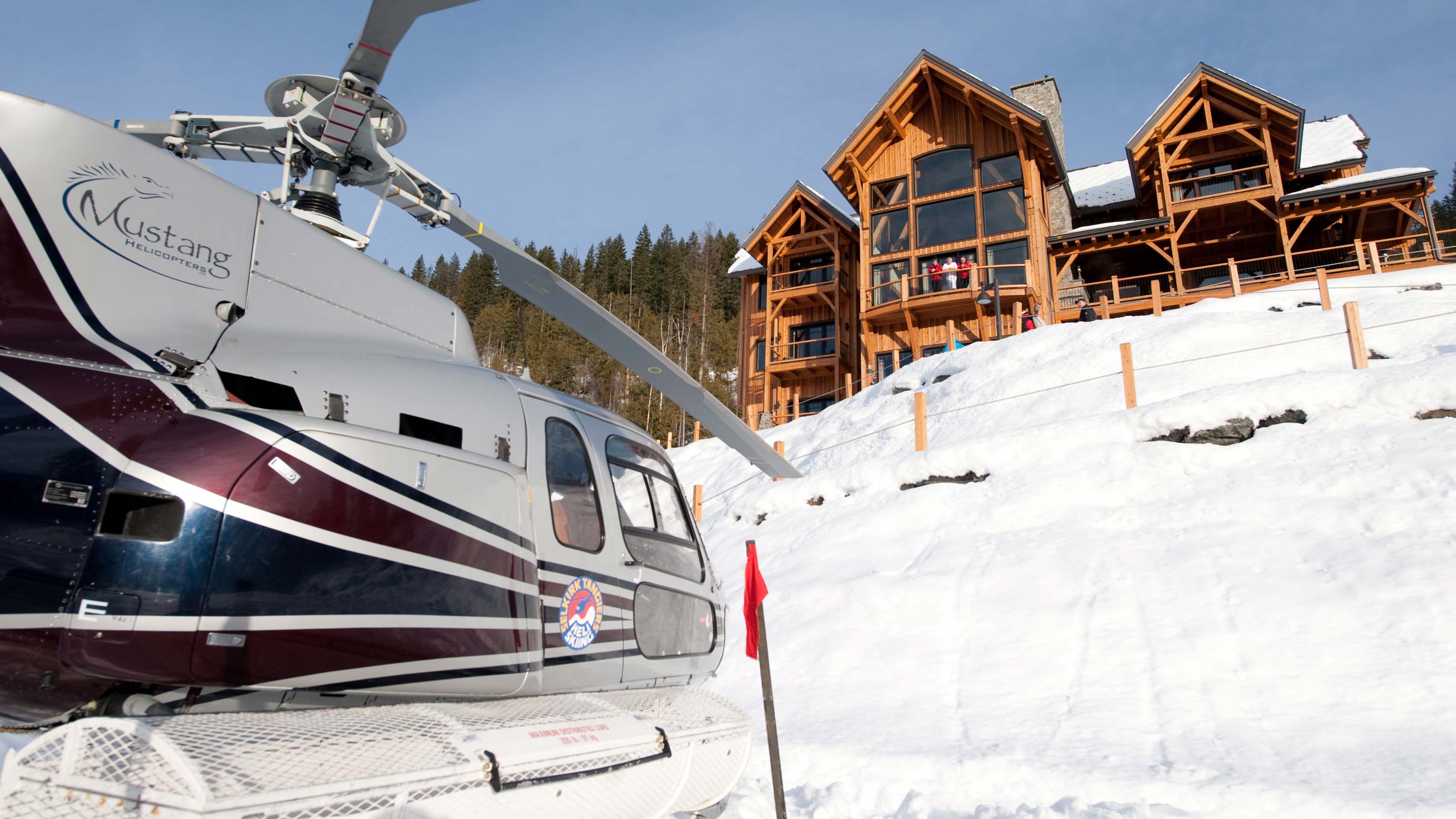 A helicopter parked in the snow with a large wooden chalet in the background. The chalet features multiple large windows and a sloped roof, surrounded by snow-covered trees.