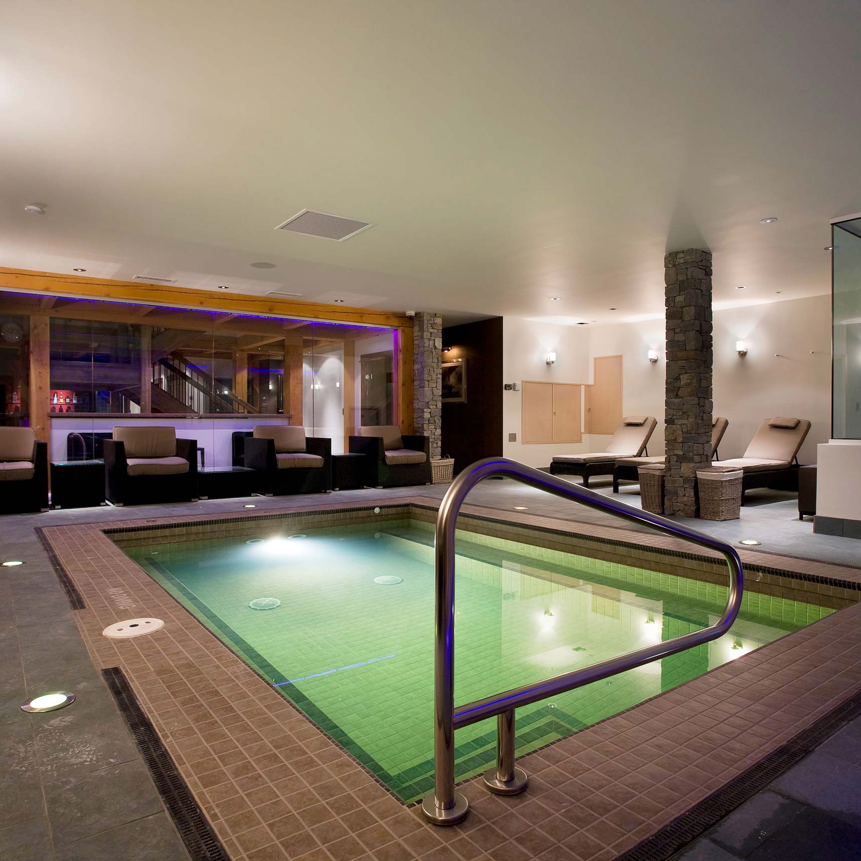 A modern spa interior featuring a small, inviting hot tub with a metal railing, surrounded by stone accents and lounge chairs. Soft ambient lighting creates a relaxing atmosphere.