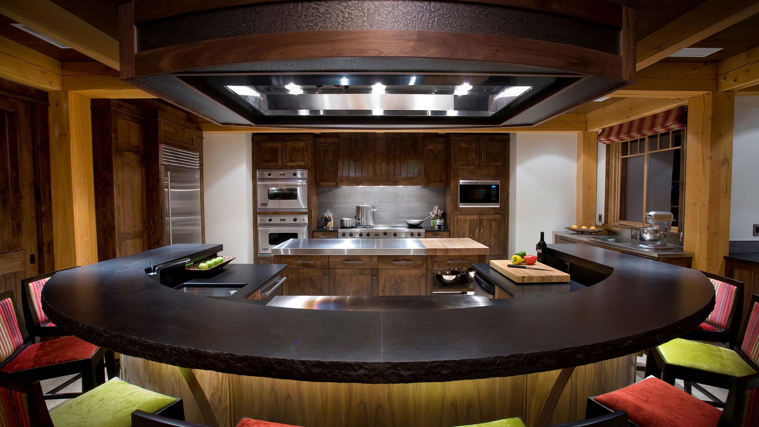 A modern kitchen featuring a curved black countertop, wooden cabinetry, stainless steel appliances, and a stylish island. Colorful bar stools are arranged around the counter, with fruits and utensils on display.
