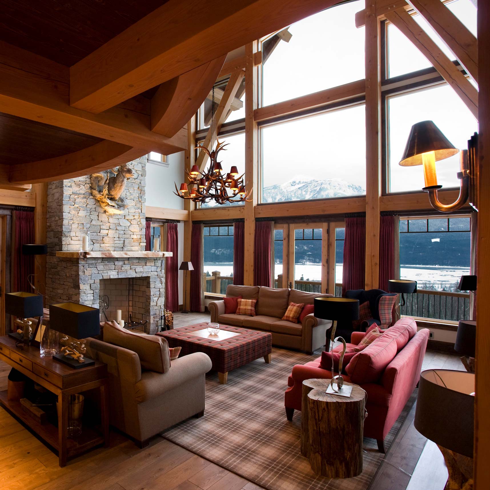 A cosy mountain lodge living room with high wooden ceilings and large windows overlooking snowy peaks. The space features a stone fireplace, plush sofas in warm tones, a wooden coffee table, and stylish lighting fixtures, creating a rustic yet inviting atmosphere.