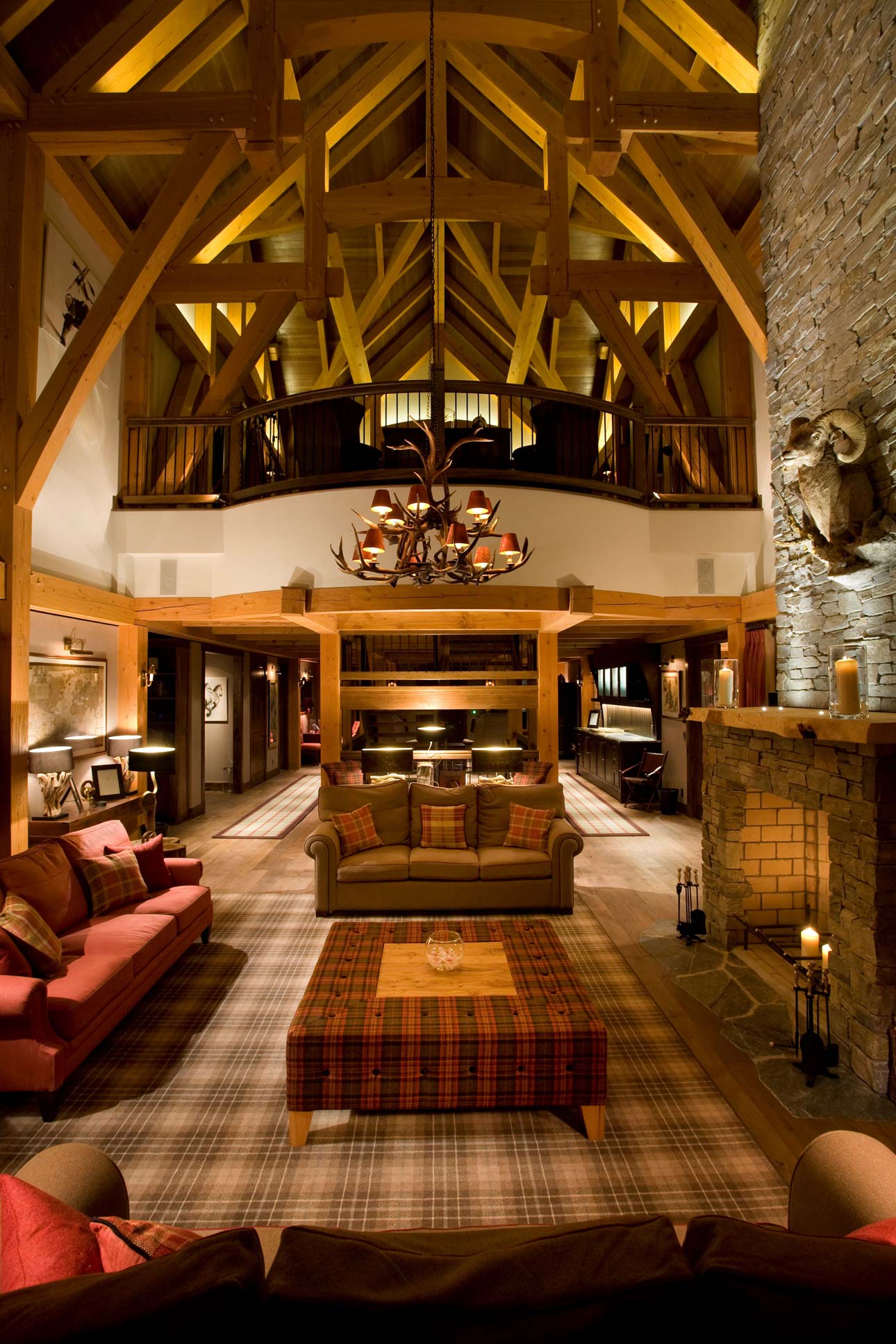 A spacious living room with high wooden ceilings and exposed beams. The room features a large stone fireplace, comfortable sofas with decorative pillows, and a central coffee table. A chandelier hangs from the ceiling, and warm lighting creates a cozy atmosphere. The floor is covered with a patterned rug, enhancing the rustic charm.