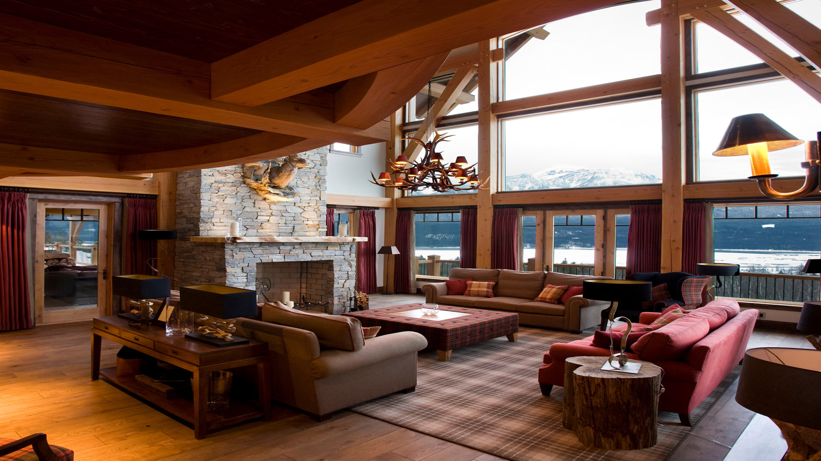 A cozy and spacious living room with large windows showcasing a mountain view. The room features wooden beams, a stone fireplace, comfortable furniture, and warm lighting. A plaid rug adds texture to the wooden floor, while a rustic side table completes the inviting atmosphere.