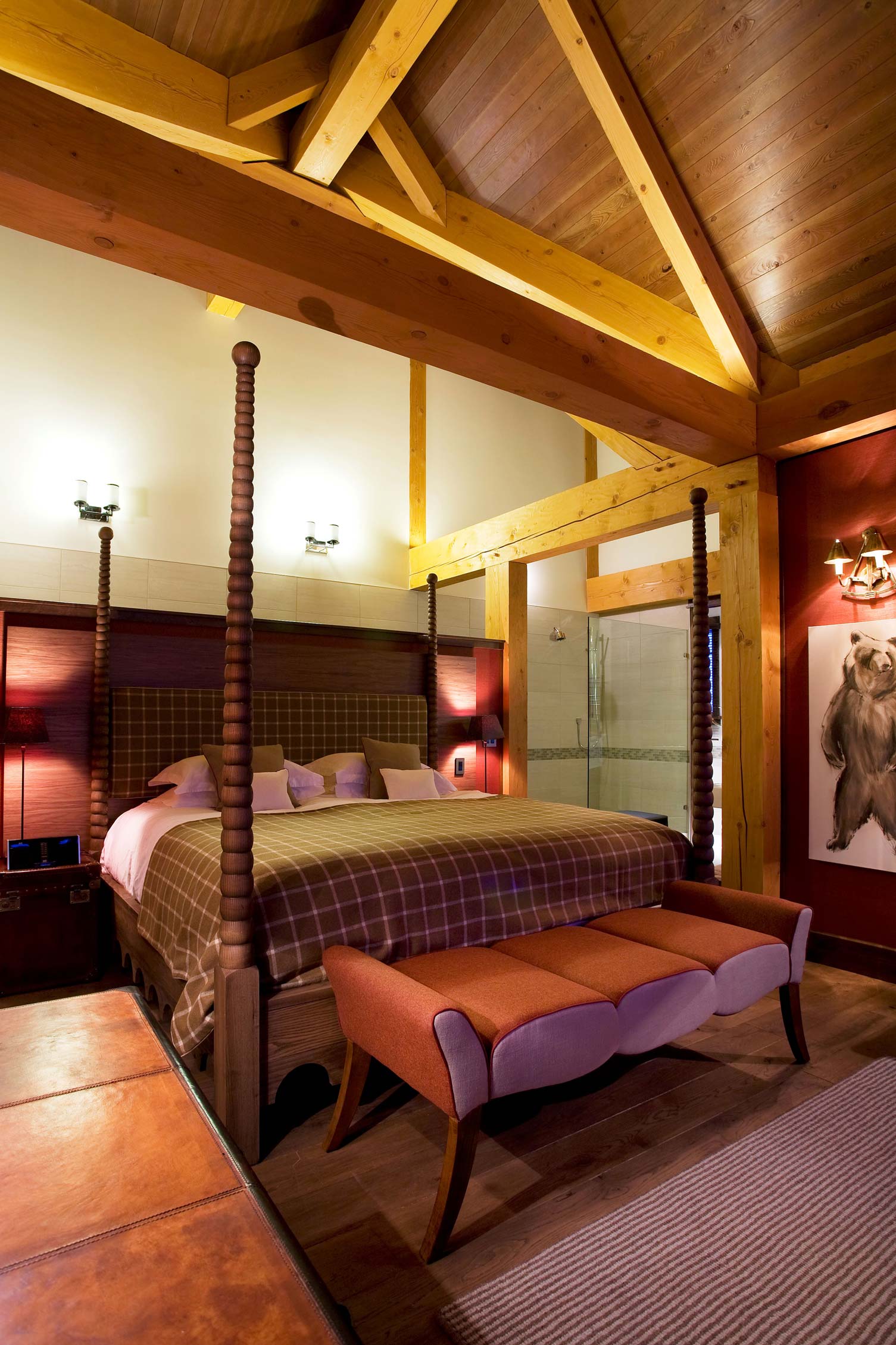 A cozy bedroom featuring a four-poster bed with a plaid duvet, a stylish bench at the foot of the bed, warm wooden beams on the ceiling, and a large bear painting on the wall. Soft lighting adds a tranquil ambiance.