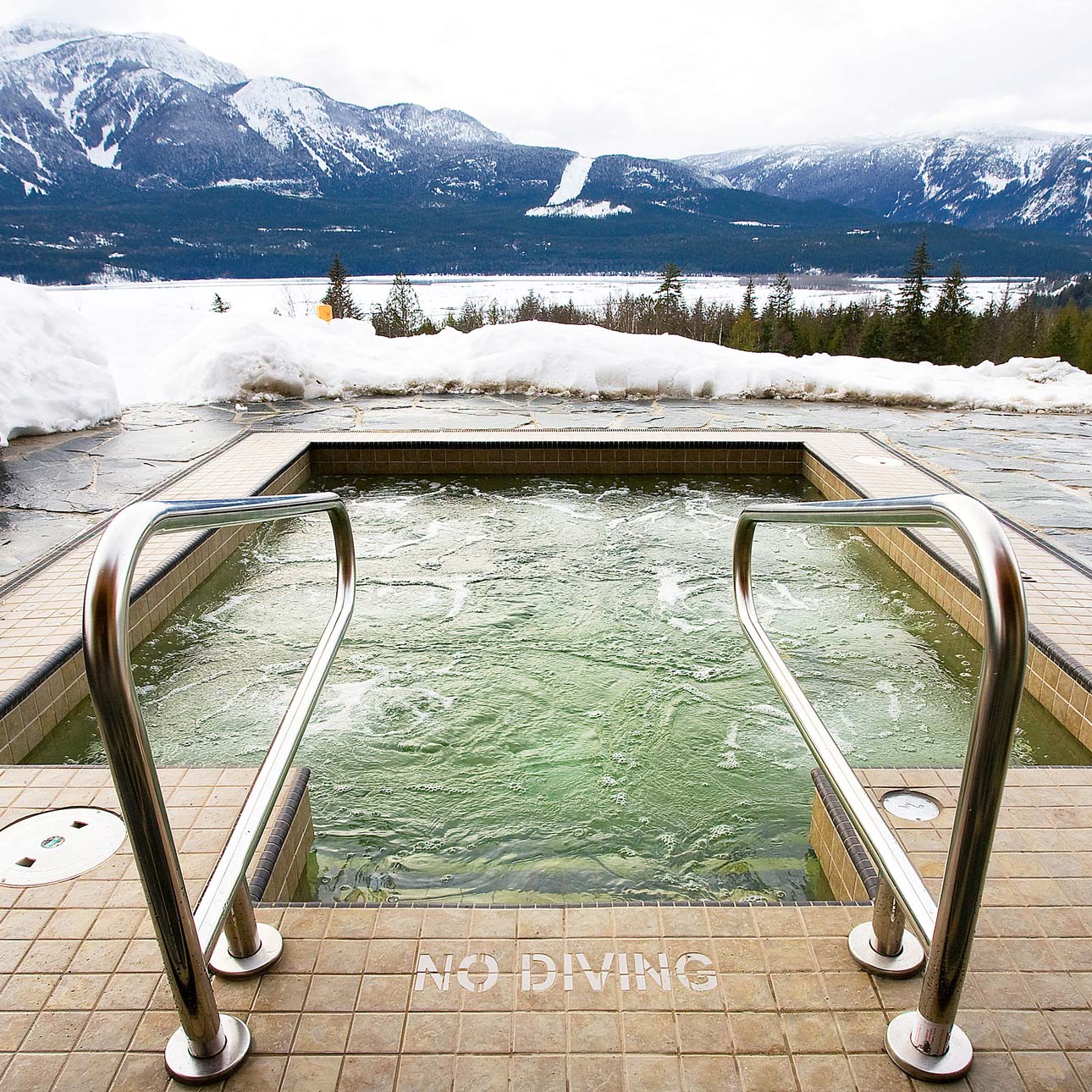 A hot tub with stainless steel railings sits on a tiled deck surrounded by snow. In the background, snow-capped mountains and a lake are visible under a cloudy sky. A "No Diving" sign is on the ground near the tub.