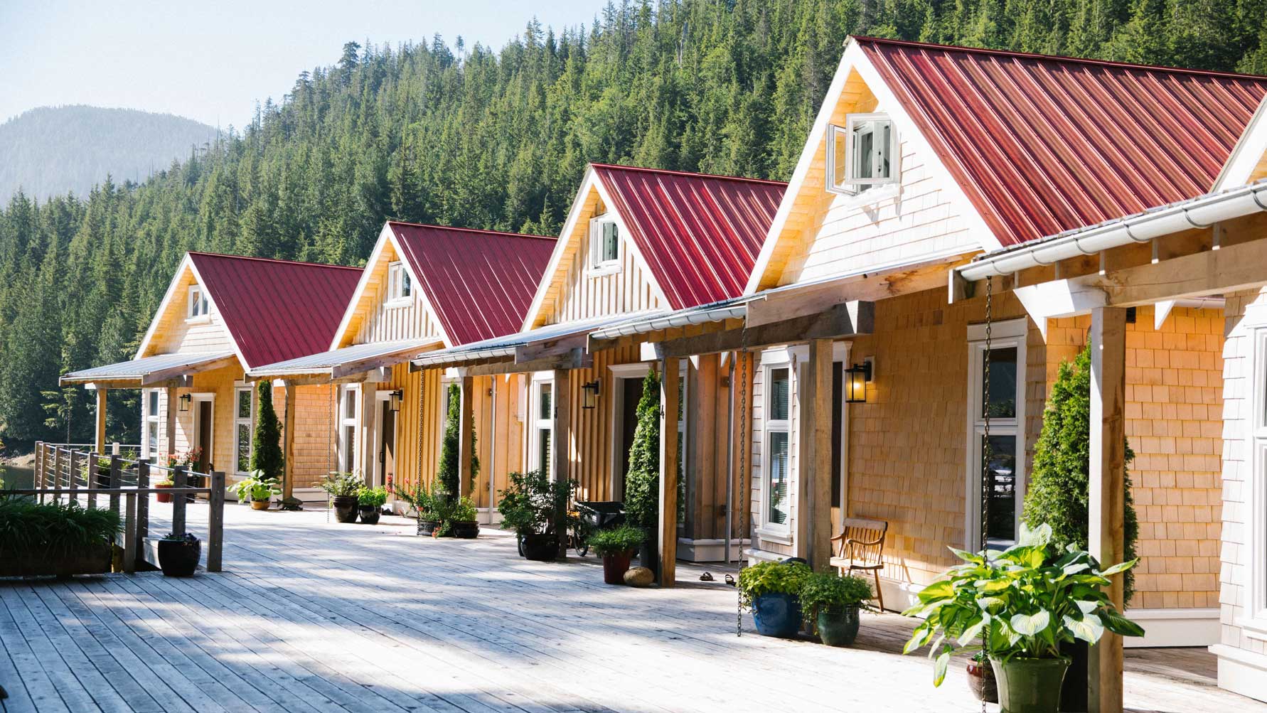 Row of charming wooden cabins with red roofs, lined up on a wooden deck, surrounded by lush green trees and mountains in the background.
