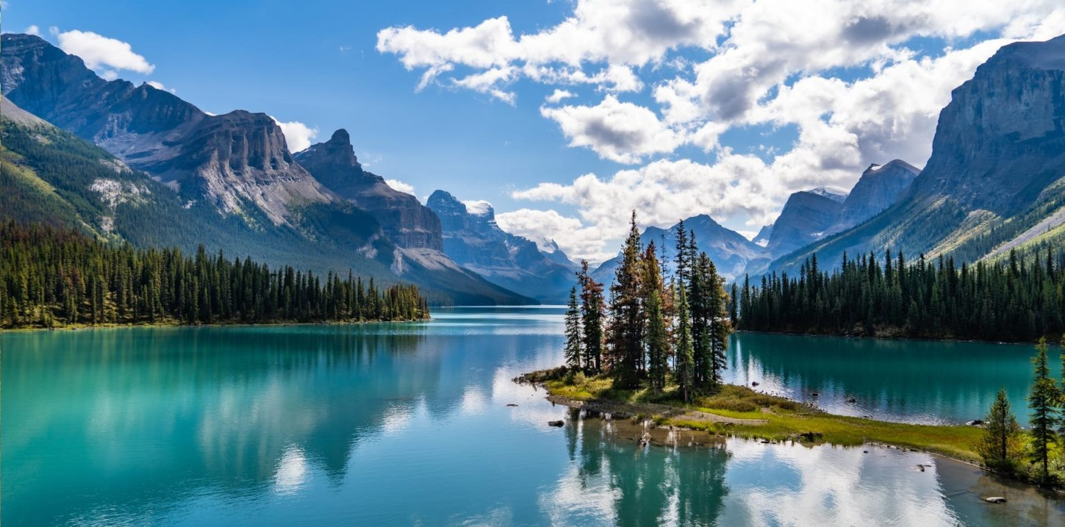 A serene lake surrounded by majestic mountains and lush greenery. The water is a vibrant turquoise, reflecting the blue sky and fluffy clouds. An island with trees is visible in the foreground.