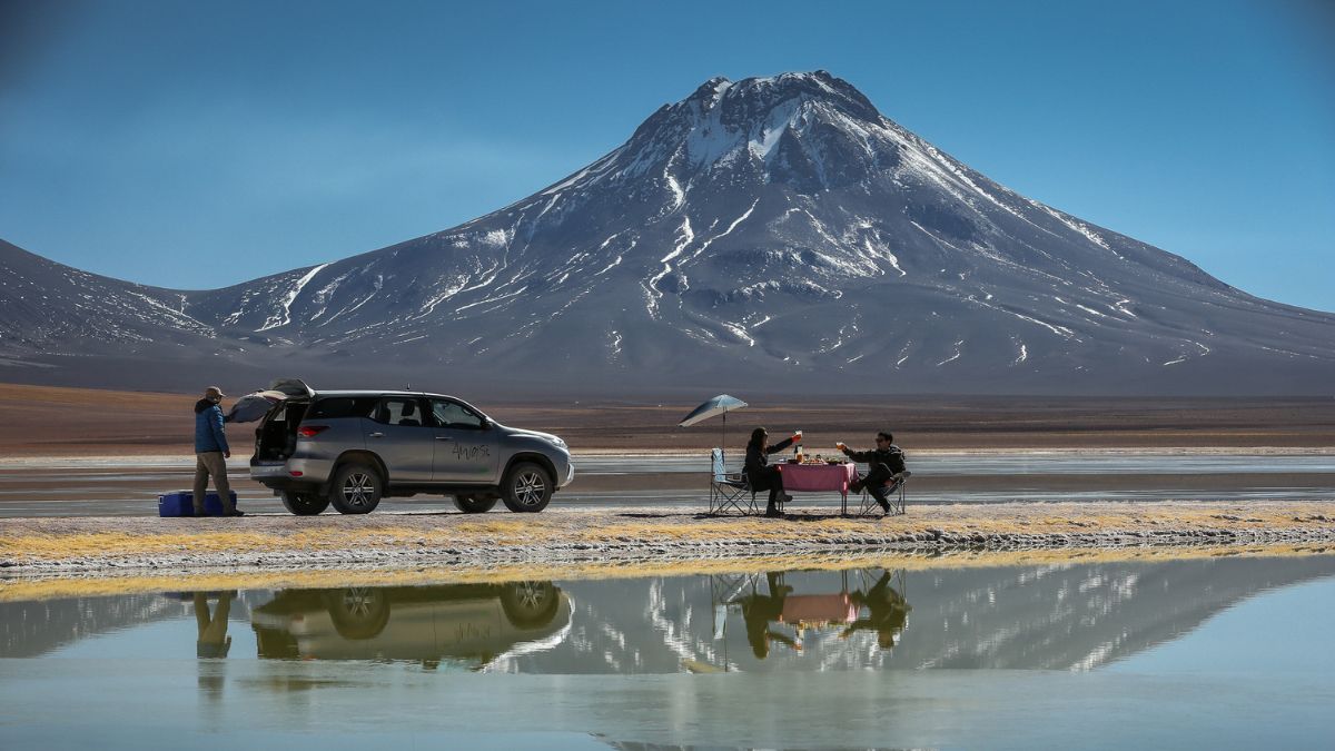 Two people having a meal at a table near a reflective salt flat with a large, snow-dusted volcano behind them.