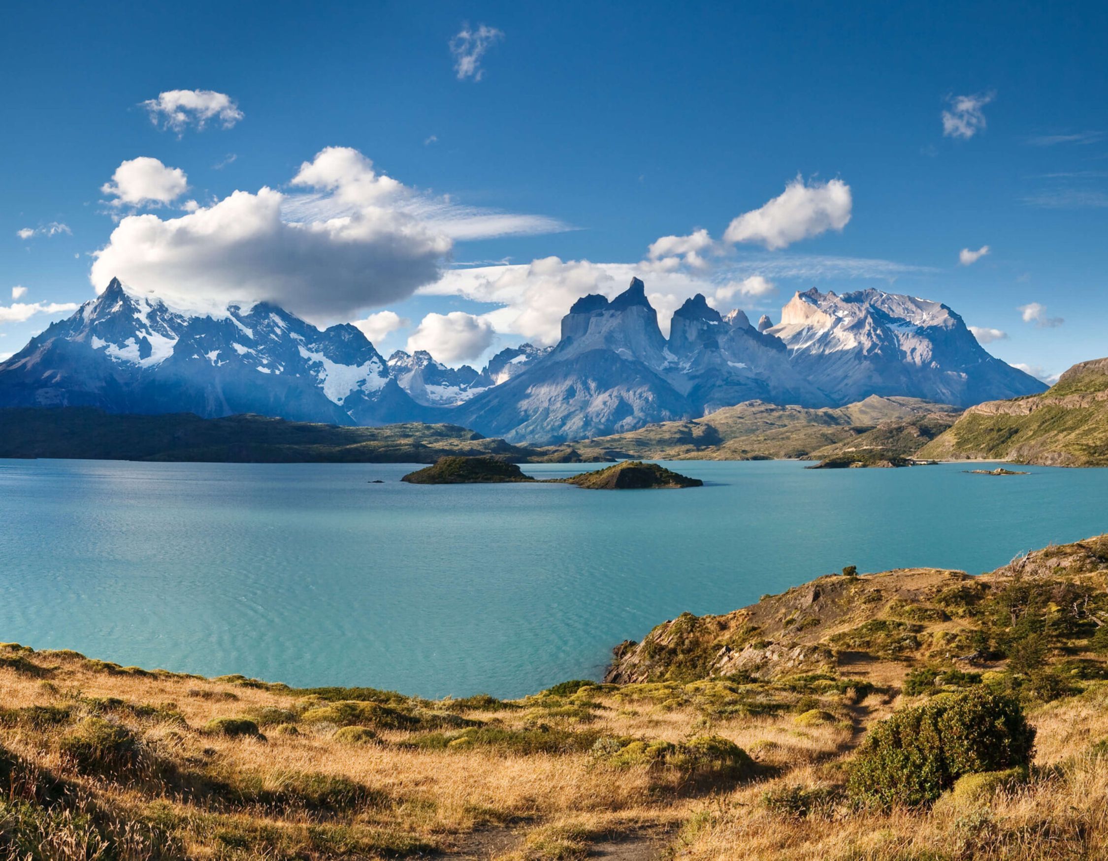 Bright blue lake surrounded by golden fields and jagged, snow-capped mountain peaks under a blue sky.