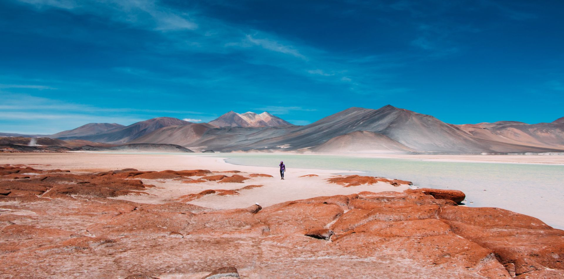 Lone hiker walking on a white salt flat surrounded by red rocks and volcanic mountains.Lone hiker walking on a white salt flat surrounded by red rocks and volcanic mountains.