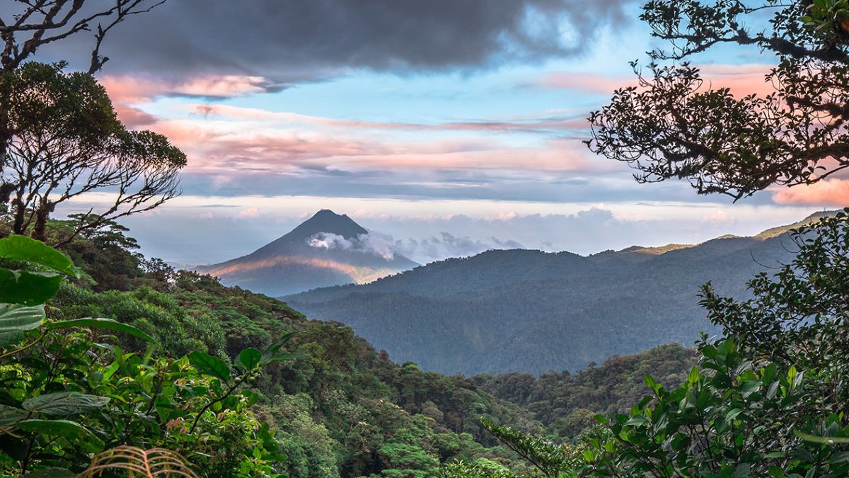 A distant, perfectly symmetrical volcanic peak rises above a dense, green tropical rainforest under a cloudy sky.