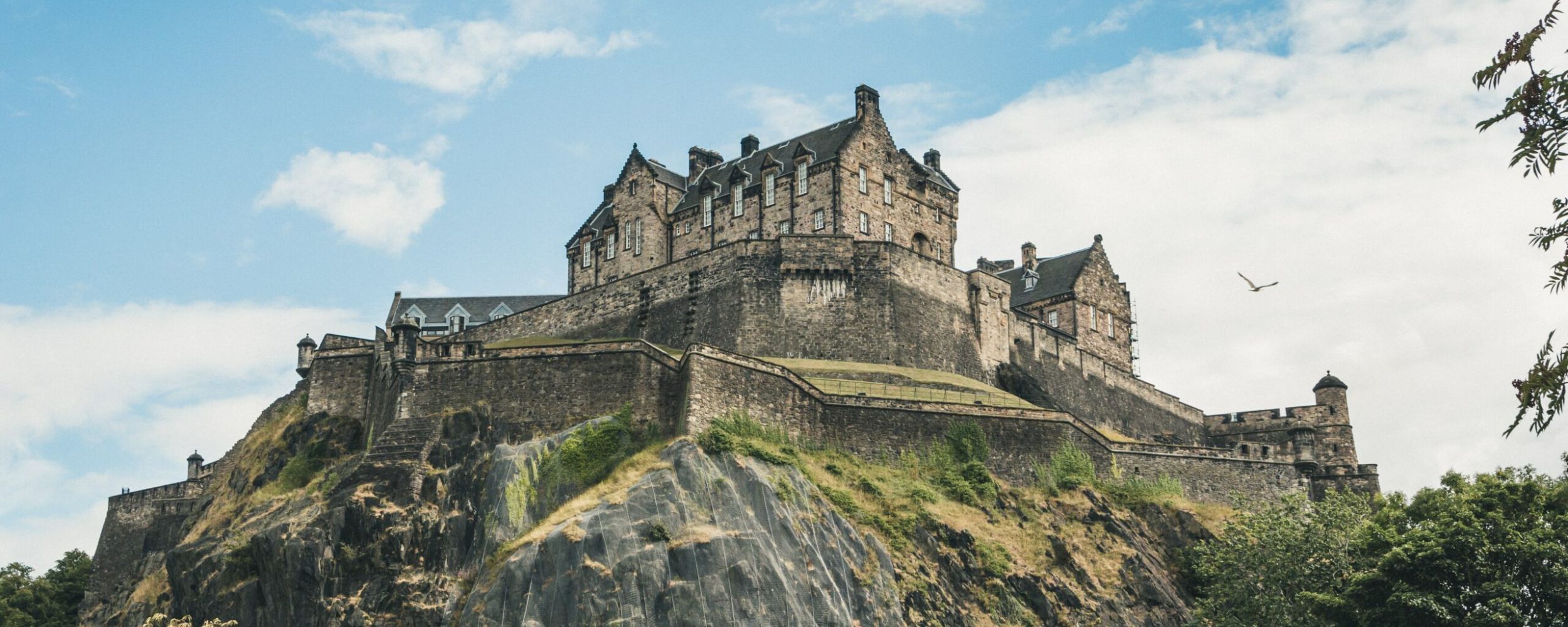 Edinburgh Castle perched atop a rugged volcanic rock overlooking green slopes and a clear sky.