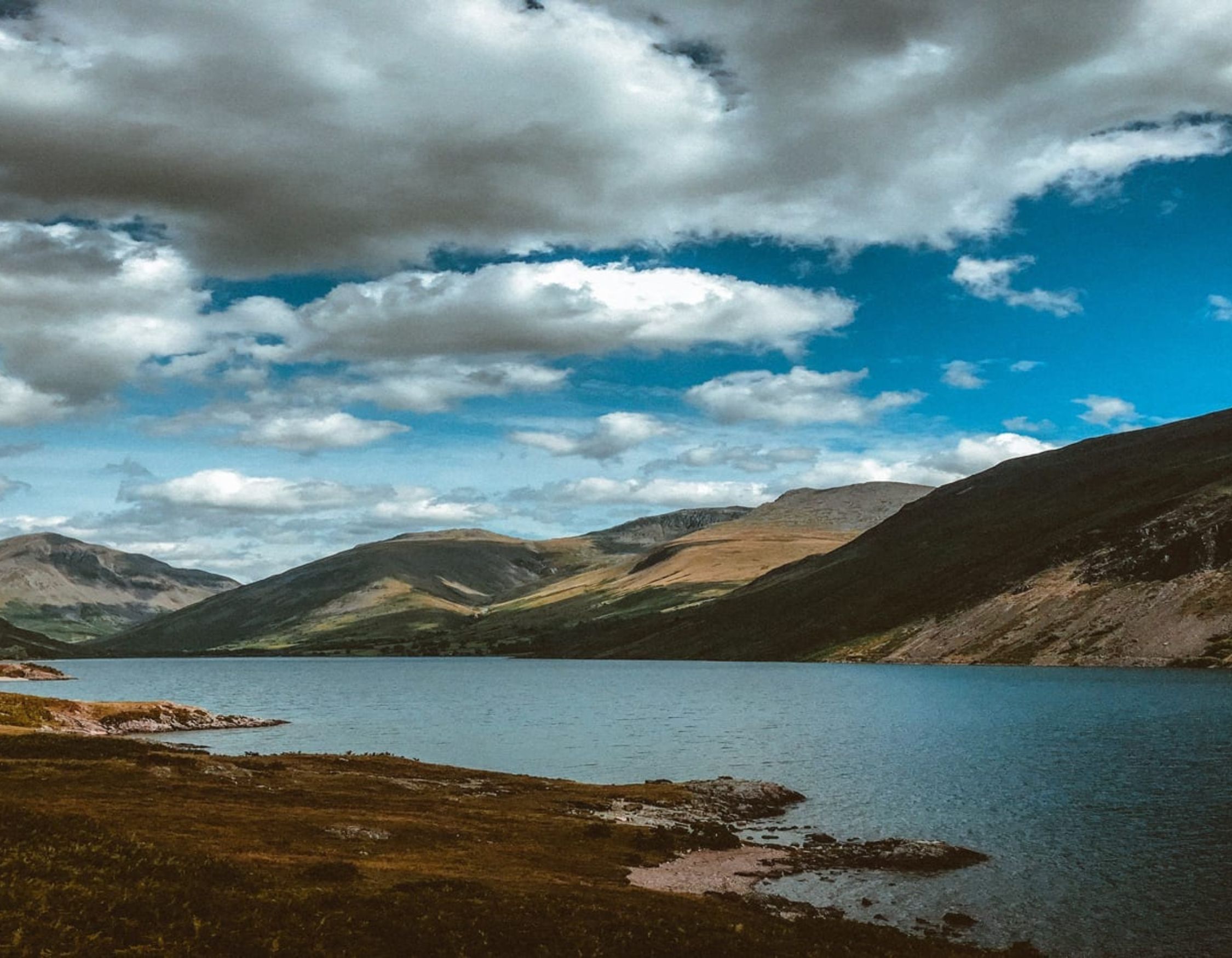 Wide view of a blue mountain lake beneath a dramatic sky filled with white and grey clouds.