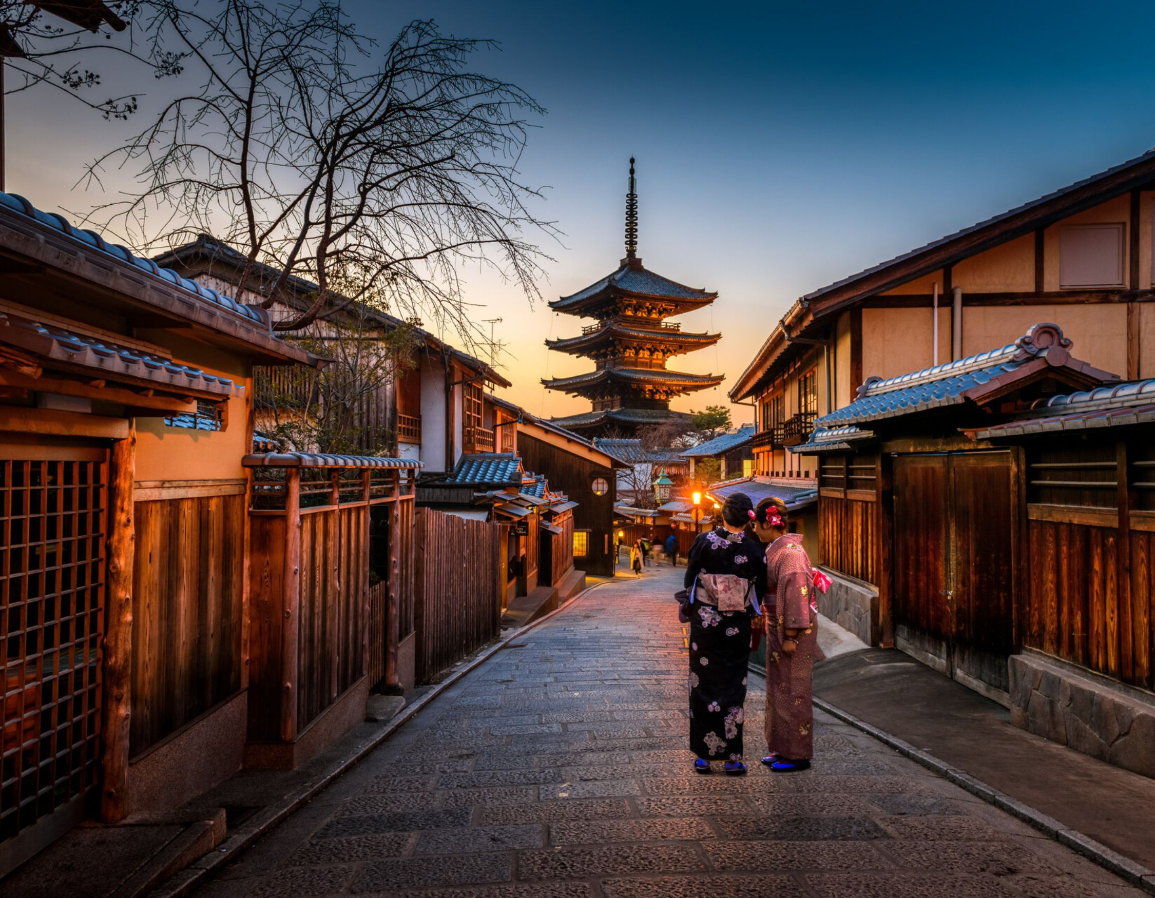 Two people in kimonos walk down a traditional Japanese street toward a tall wooden pagoda at dusk.