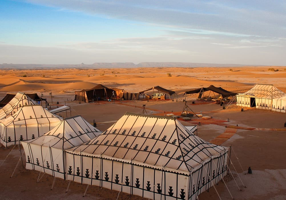 Luxury desert camp featuring several white and black patterned tents under a pale blue sky.