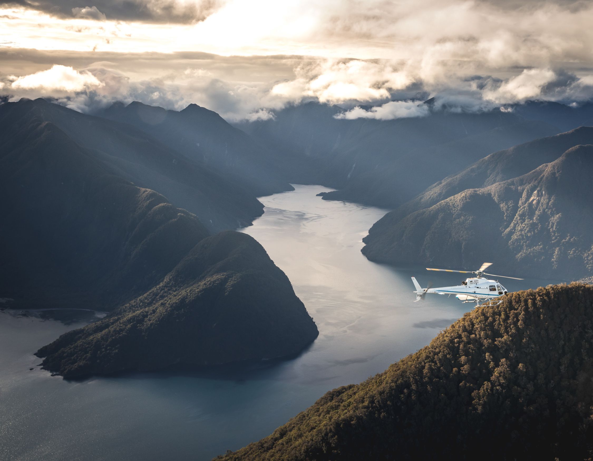 High-angle view of a white helicopter flying over a forested ridge toward a wide river valley nestled in mountains.