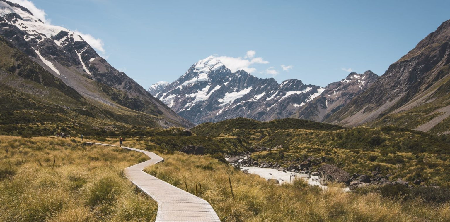 A winding wooden boardwalk leads through a grassy valley toward a massive, snow-covered mountain peak.