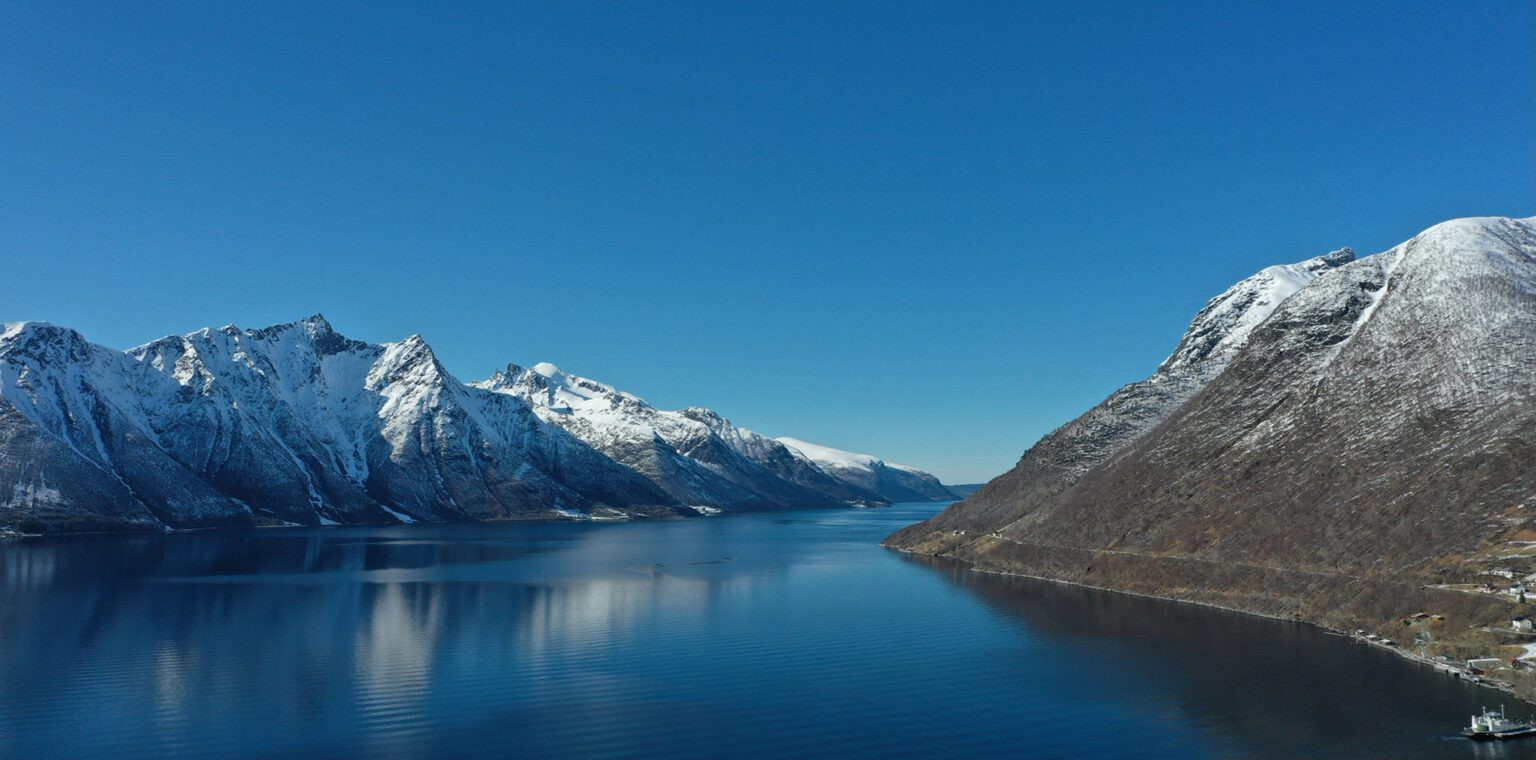 A wide, calm blue fjord reflects the snow-capped mountain peaks lining the water under a clear sky.