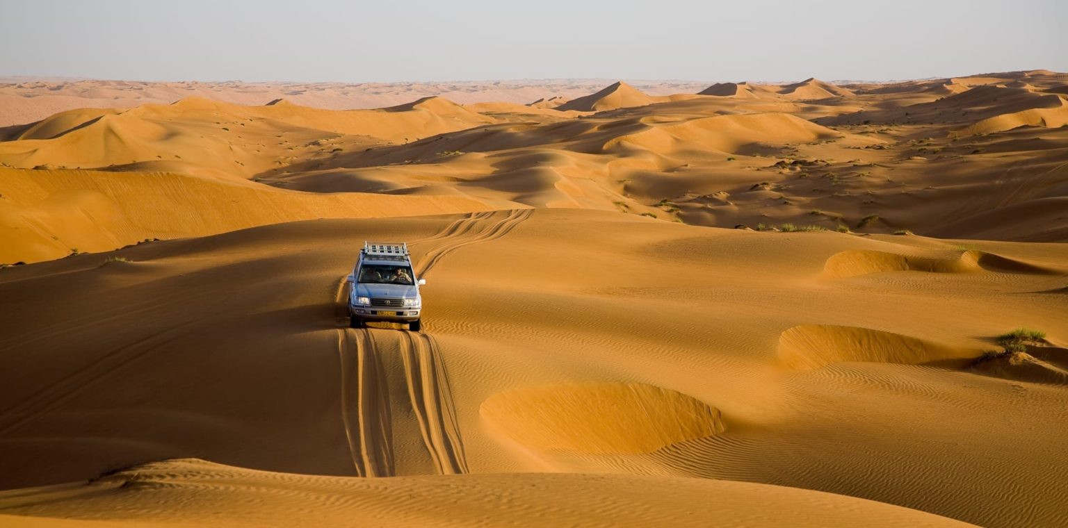 Silver SUV driving through rolling sand dunes in a vast desert, leaving tire tracks in the sand.